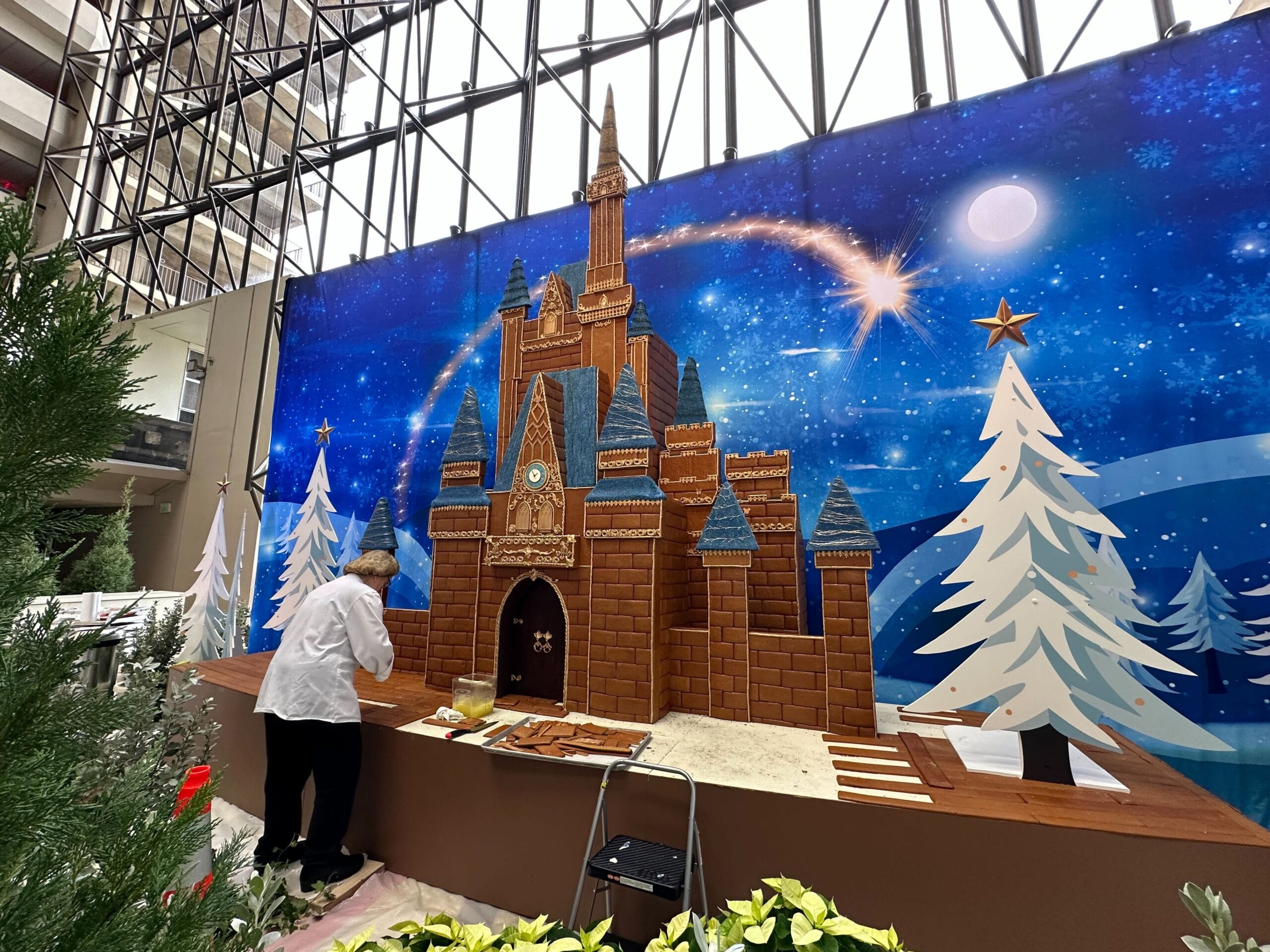 Person decorating a large gingerbread castle display with snowy trees and a starry backdrop indoors.