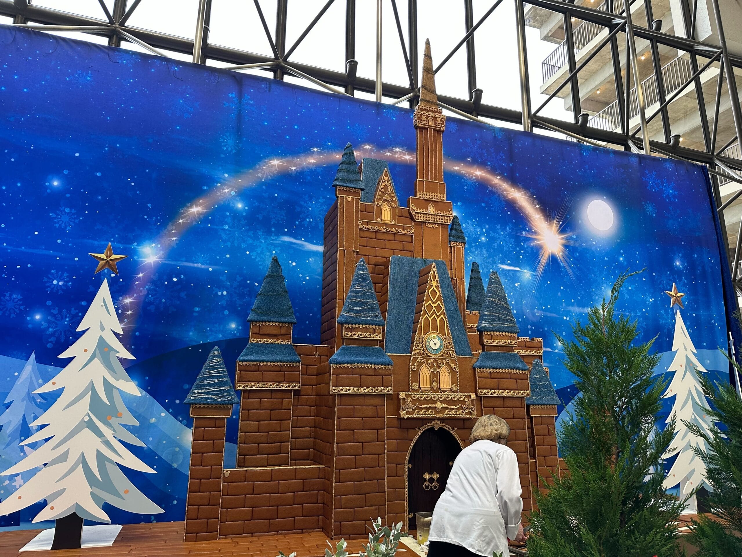 Person decorating a large gingerbread castle display with Christmas trees in front of a backdrop featuring a starry night sky.
