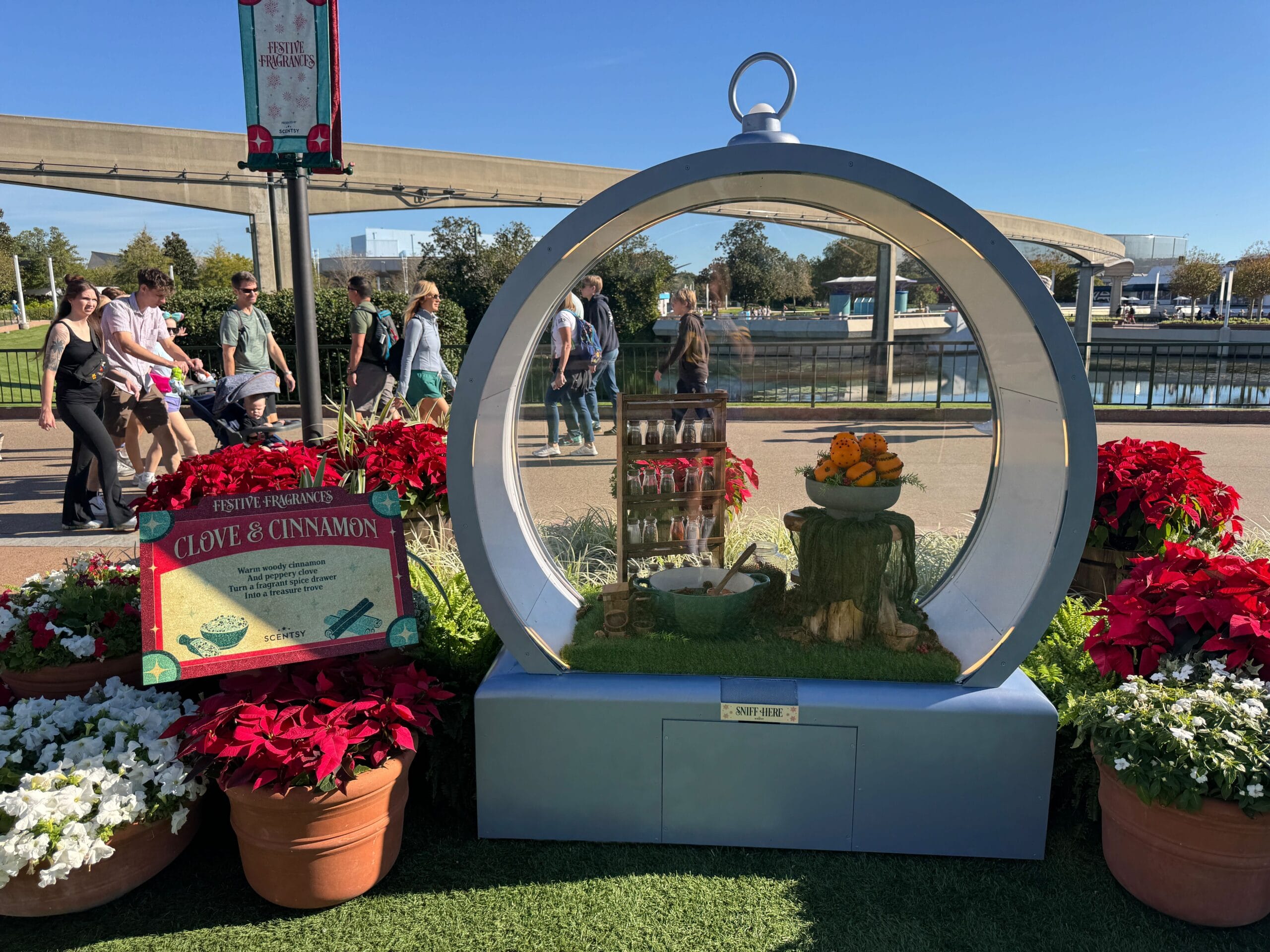 A circular display featuring a Clove & Cinnamon theme, surrounded by potted red poinsettias and white flowers, sets the scene at the EPCOT International Festival of the Holidays. People walk by under a clear blue sky, enchanted by the festive fragrances wafting through the air.