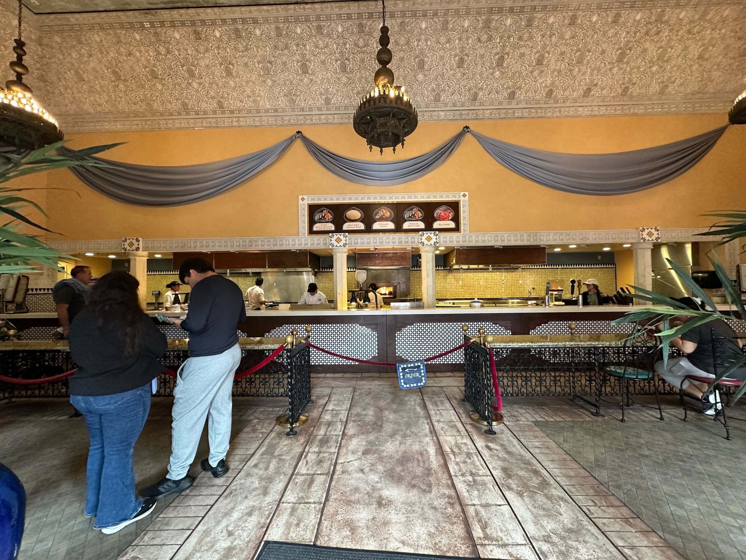 People stand in line at Tangierine Cafe: Flavors of the Medina, where the restaurant counter is adorned with decorative drapes and ornate lighting, all set against a yellow and beige color scheme.
