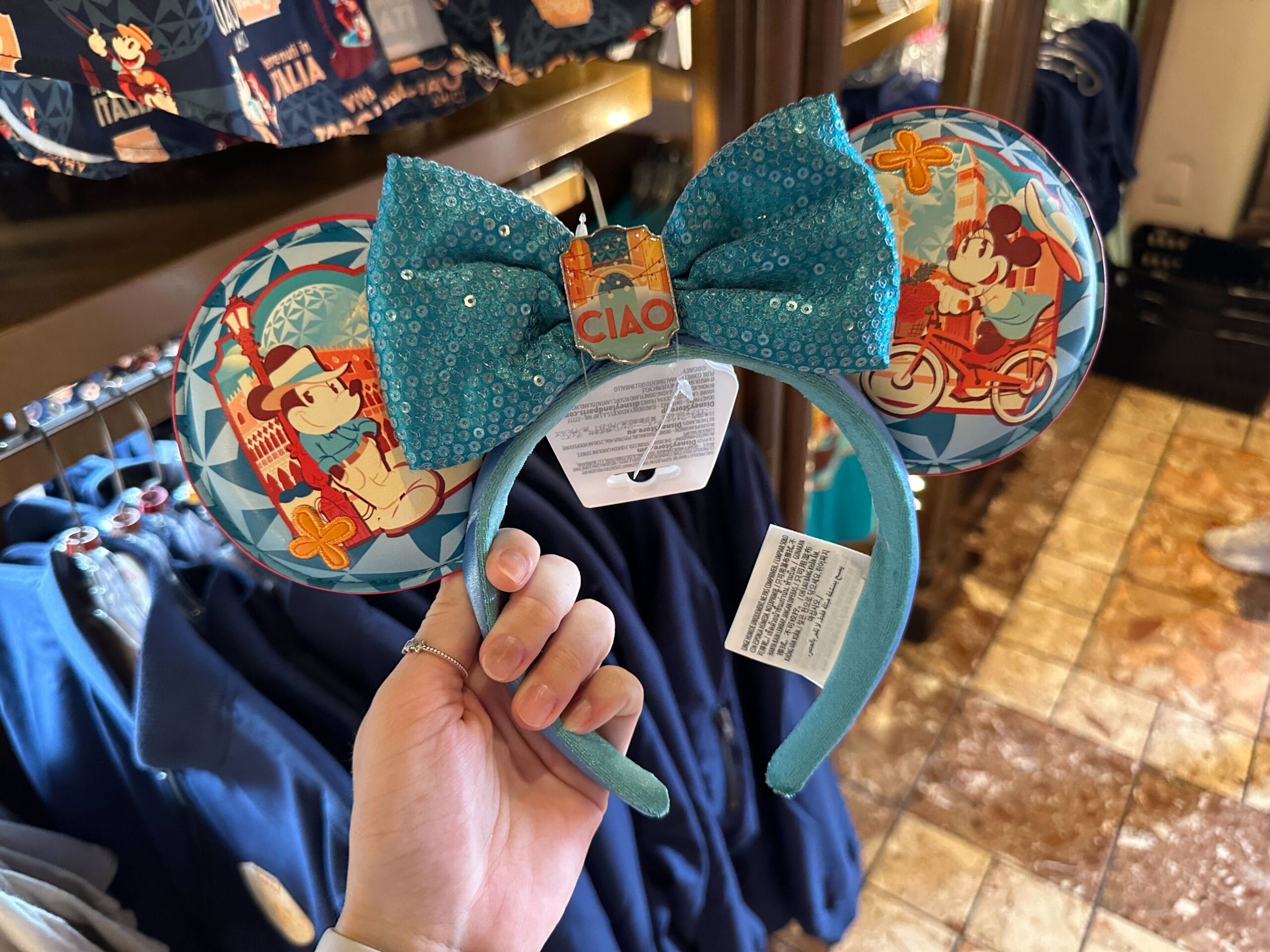 A hand holds a blue headband featuring large ears with cartoon designs and a central sequin bow, with a tab labeled "CIAO.