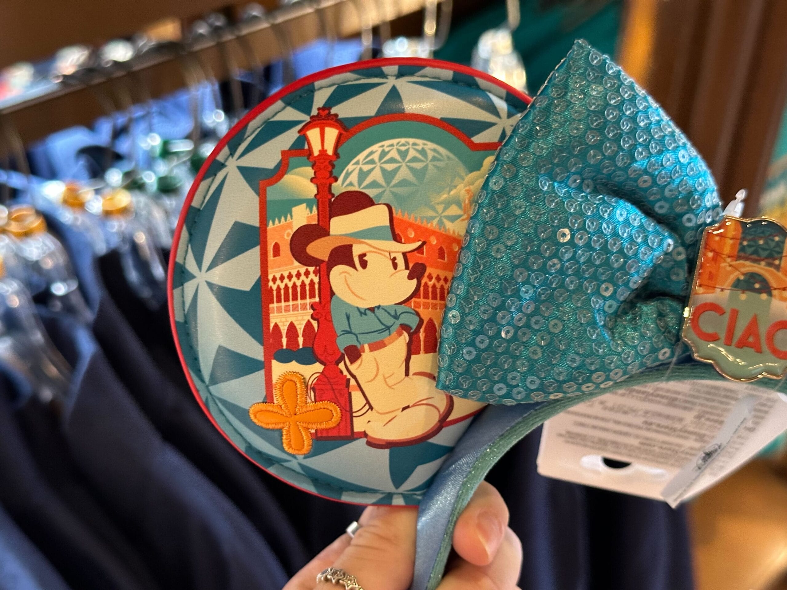 A hand holds a Mickey Mouse themed decorative coaster with a blue bow. Mickey is wearing a hat and scarf, with a backdrop of an architectural scene.