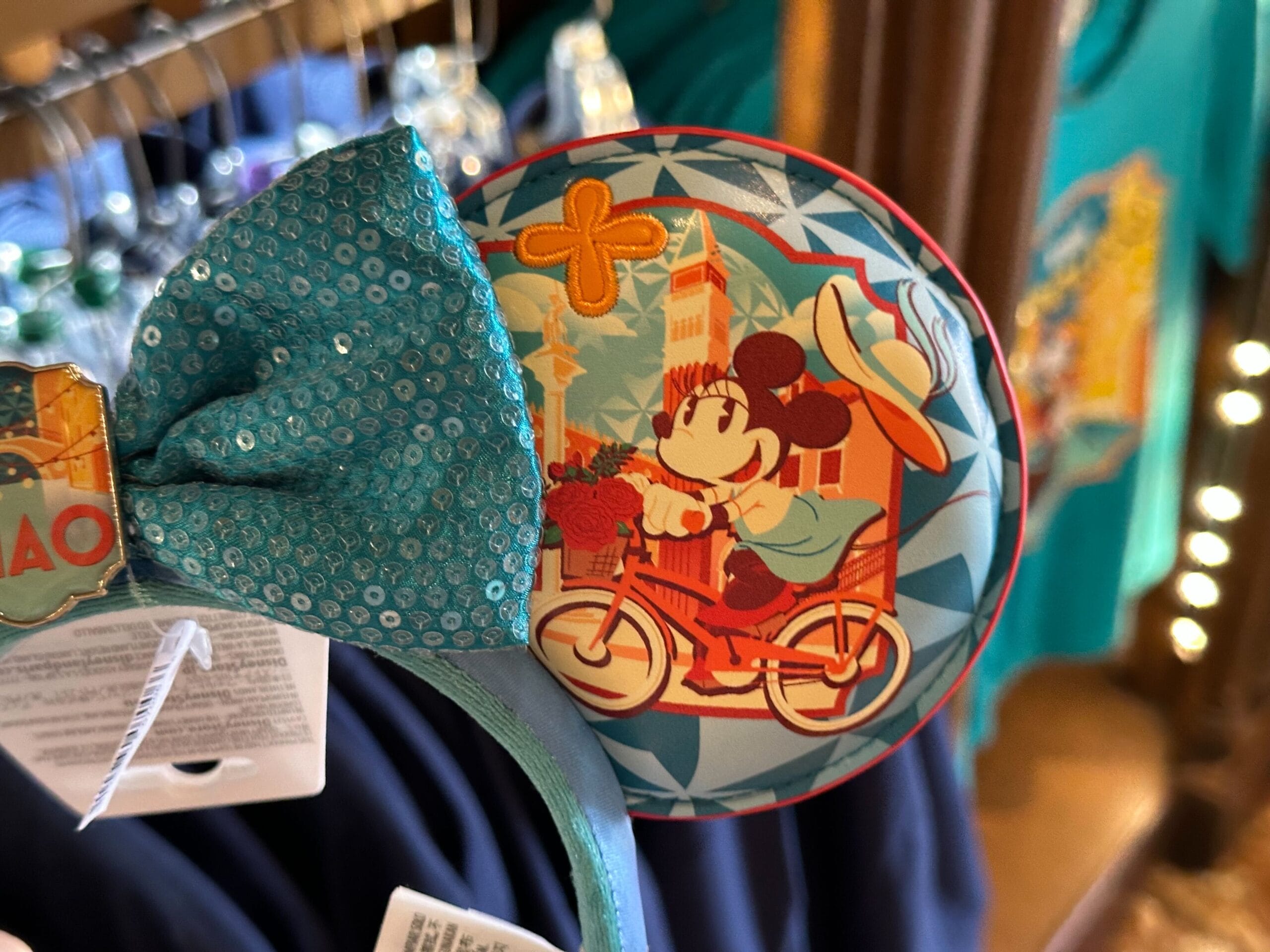 Close-up of a teal bow headband featuring a decorative round pin with a cartoon mouse riding a bicycle in front of a building and orange flowers.