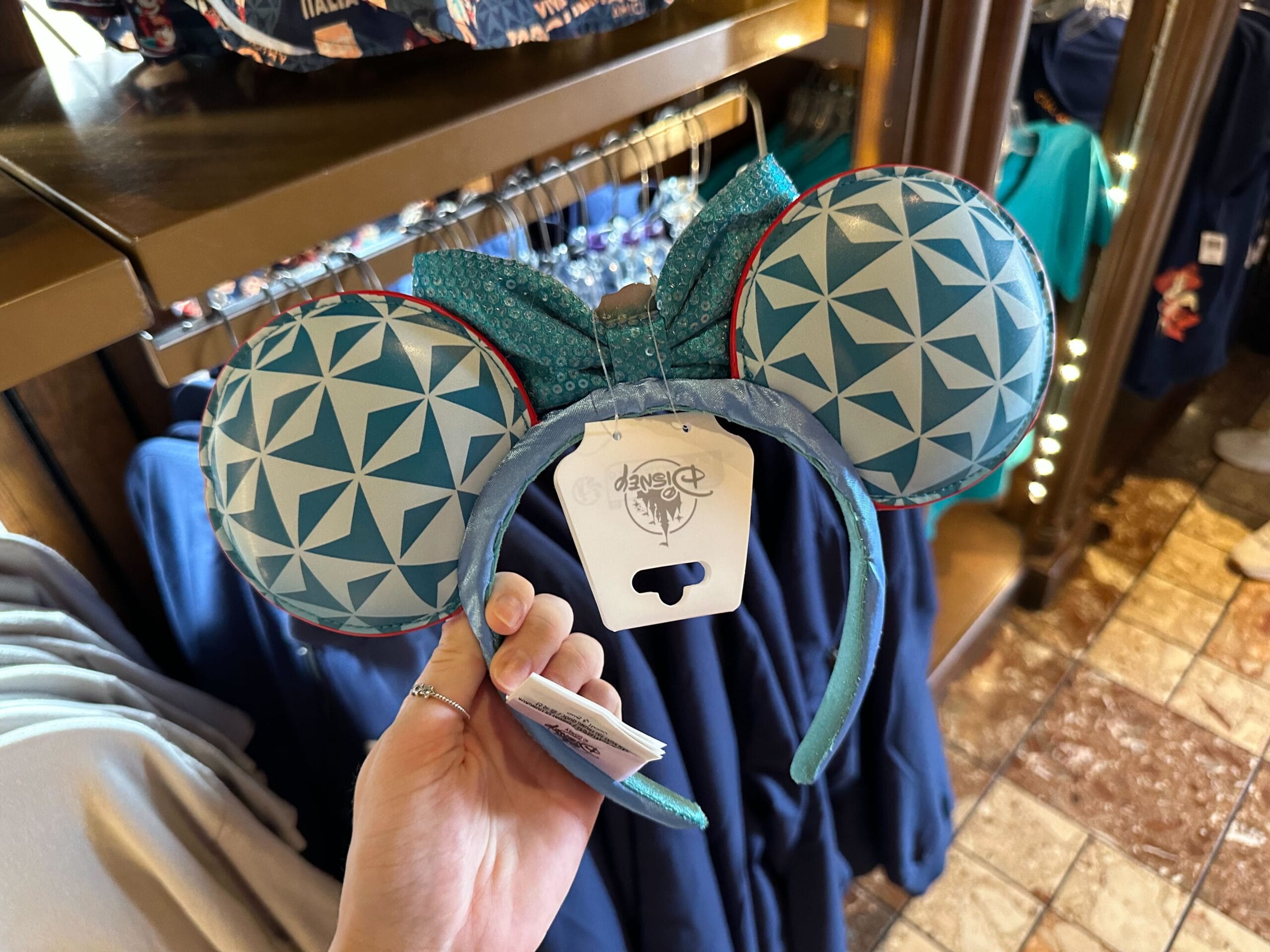 A person holds a pair of Disney-themed blue mouse ears with geometric patterns and a bow, displayed in front of clothing in a store setting.