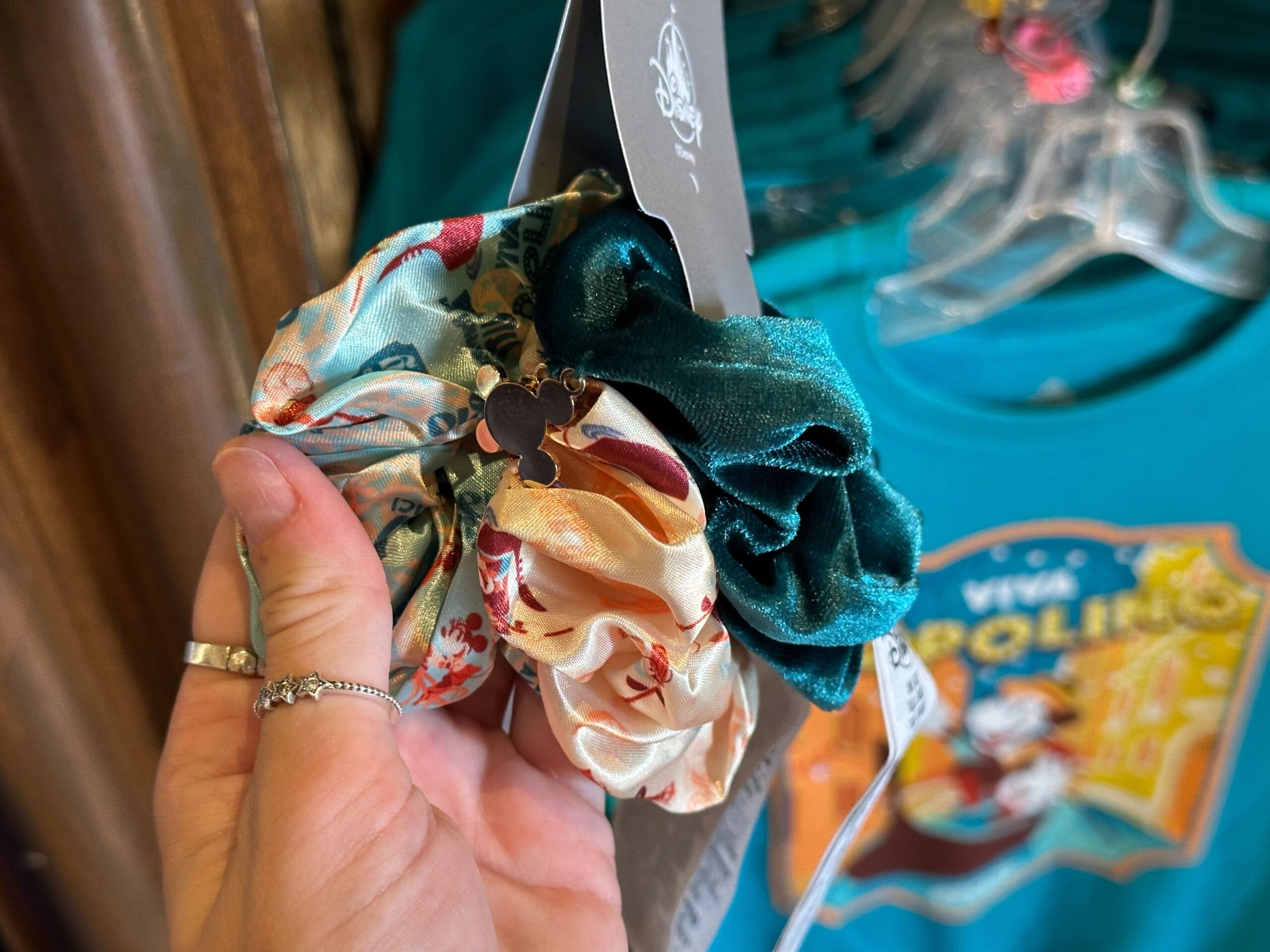 A hand holding three scrunchies: one floral, one cream, and one green velvet. A keychain with a silhouette of a certain character is attached. Background features a colorful t-shirt on a rack.