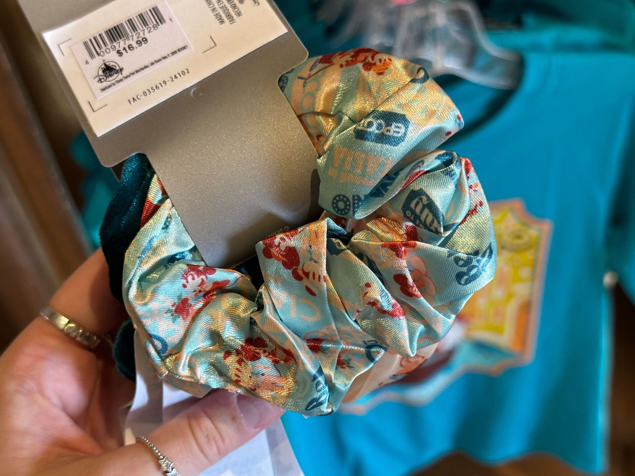 Close-up of a hand holding scrunchies with colorful patterns, a price tag attached. A teal shirt with a graphic design is visible in the background.