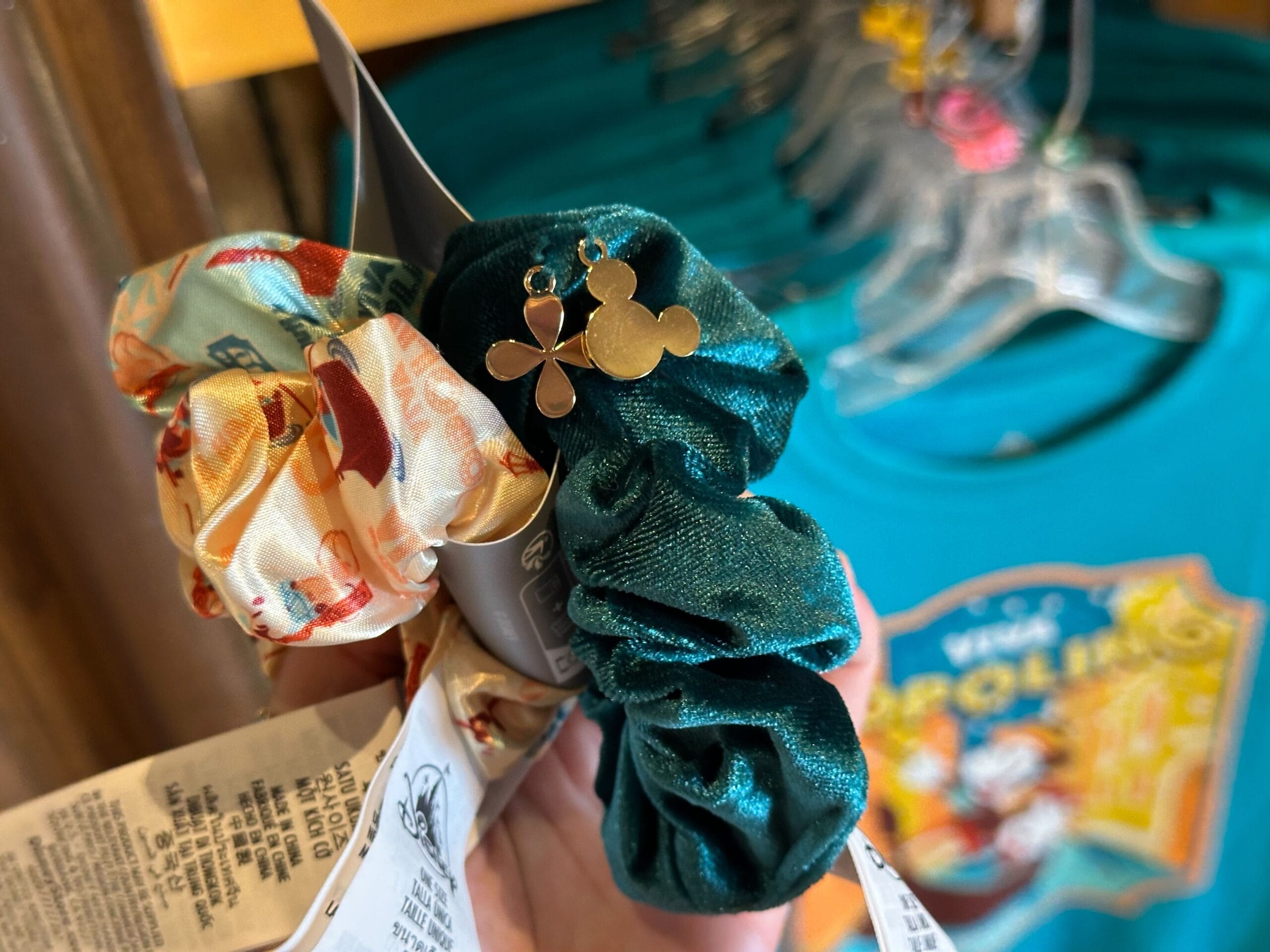 A hand holds two scrunchies: one floral and one green. The green scrunchie has a gold Mickey Mouse-shaped charm. A graphic T-shirt is visible in the background.