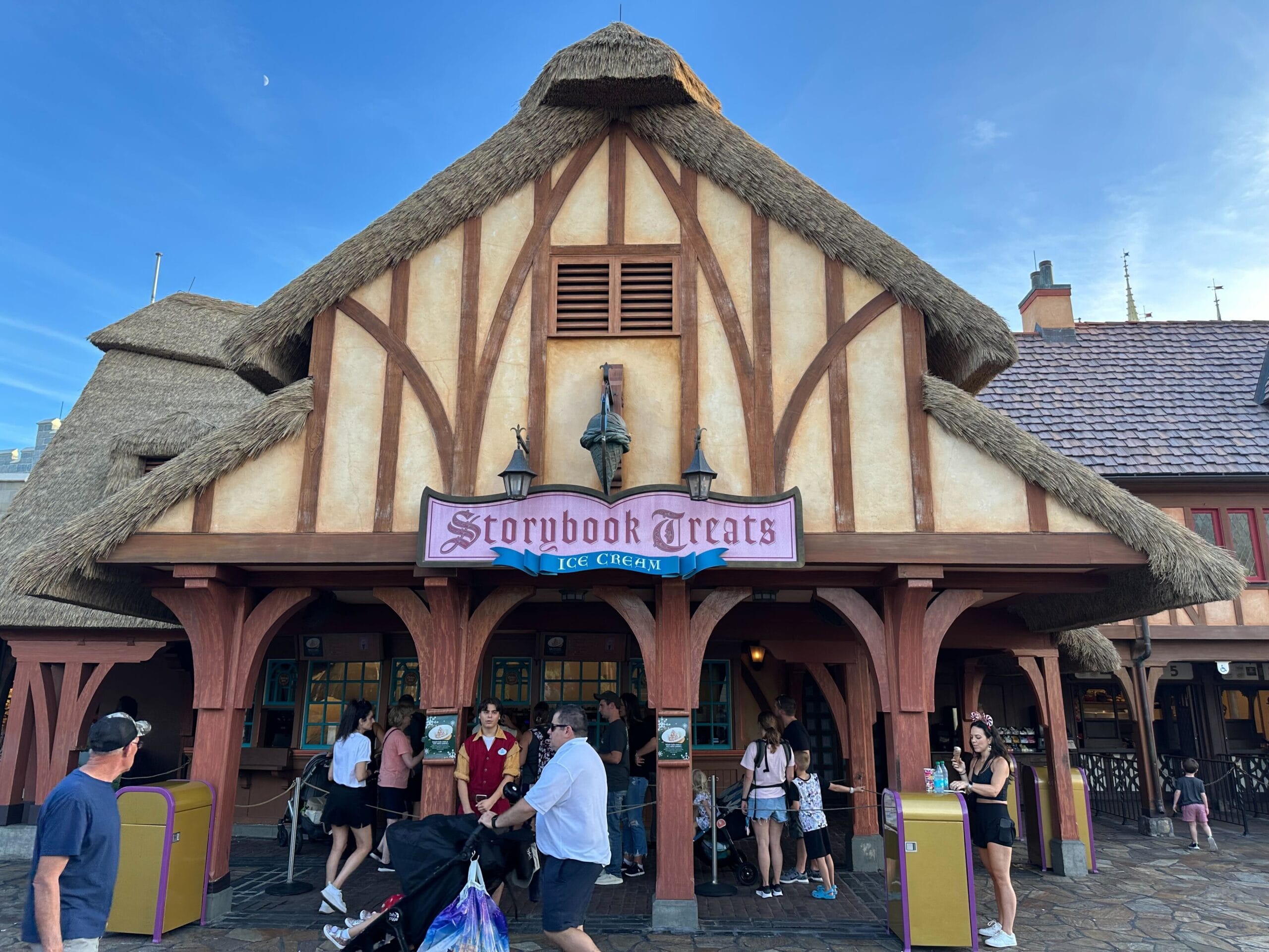 People gather outside the Storybook Treats ice cream shop, a rustic building with a thatched roof and wooden beams, on a sunny day, eager to sample the special Christmas treats at Magic Kingdom.