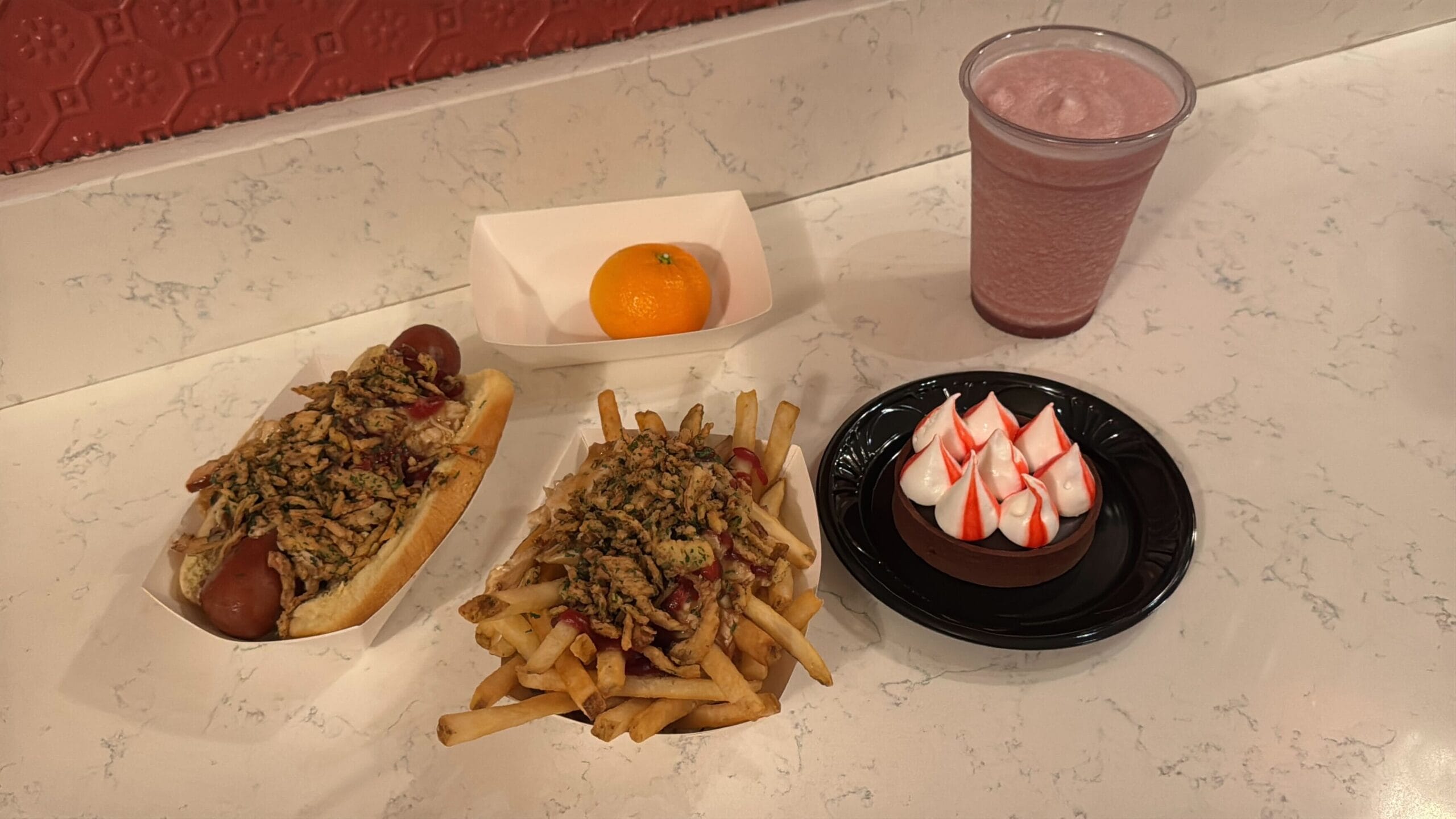 At Casey's Corner, enjoy a hot dog with crispy toppings, loaded fries, a juicy orange, a round dessert with red and white swirls, and a pink milkshake—all perfectly displayed on a marble counter.