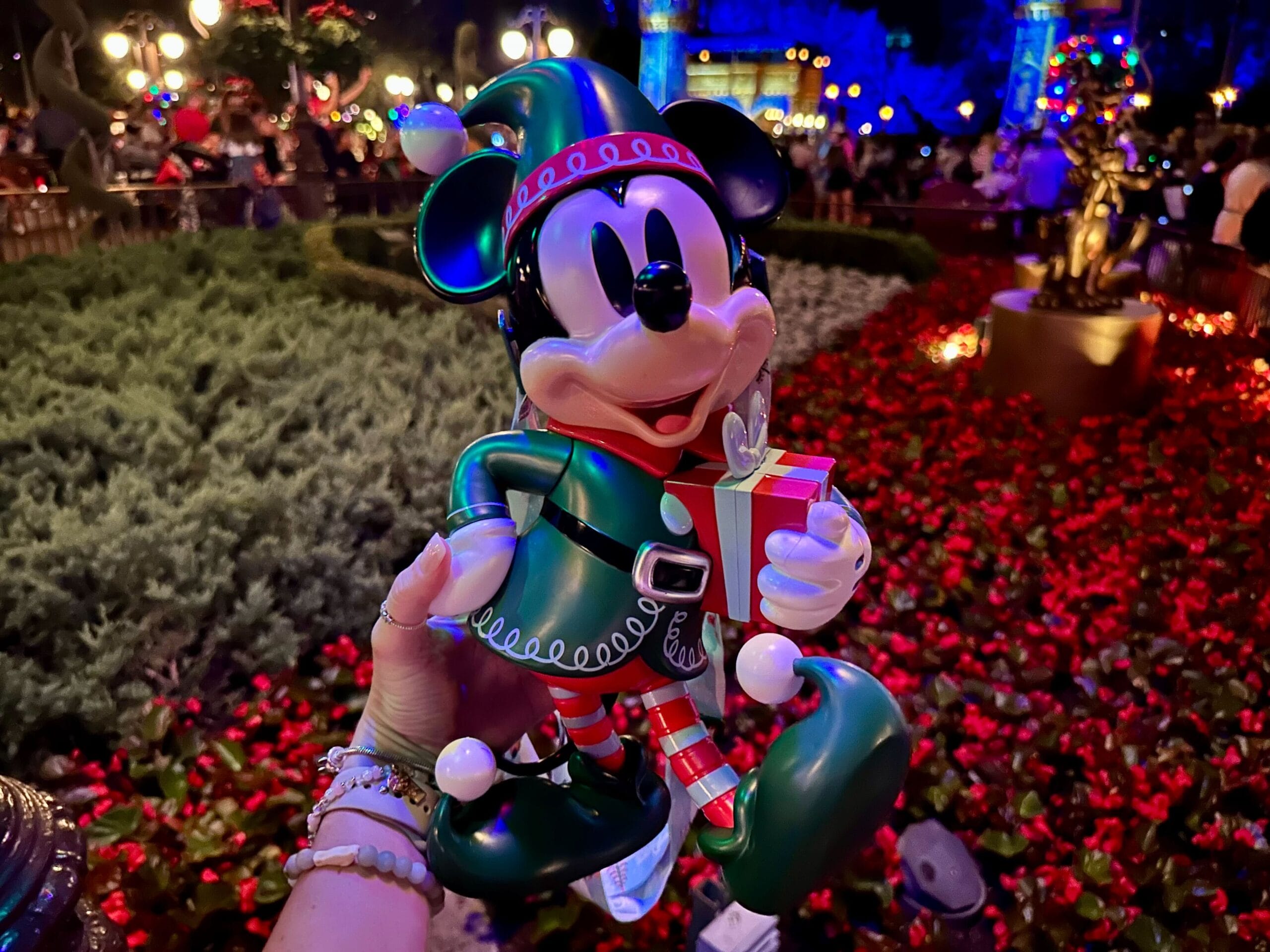 A hand holds a Christmas-themed Mickey Mouse figurine dressed as an elf, in front of a garden with colorful flowers and festive lights.