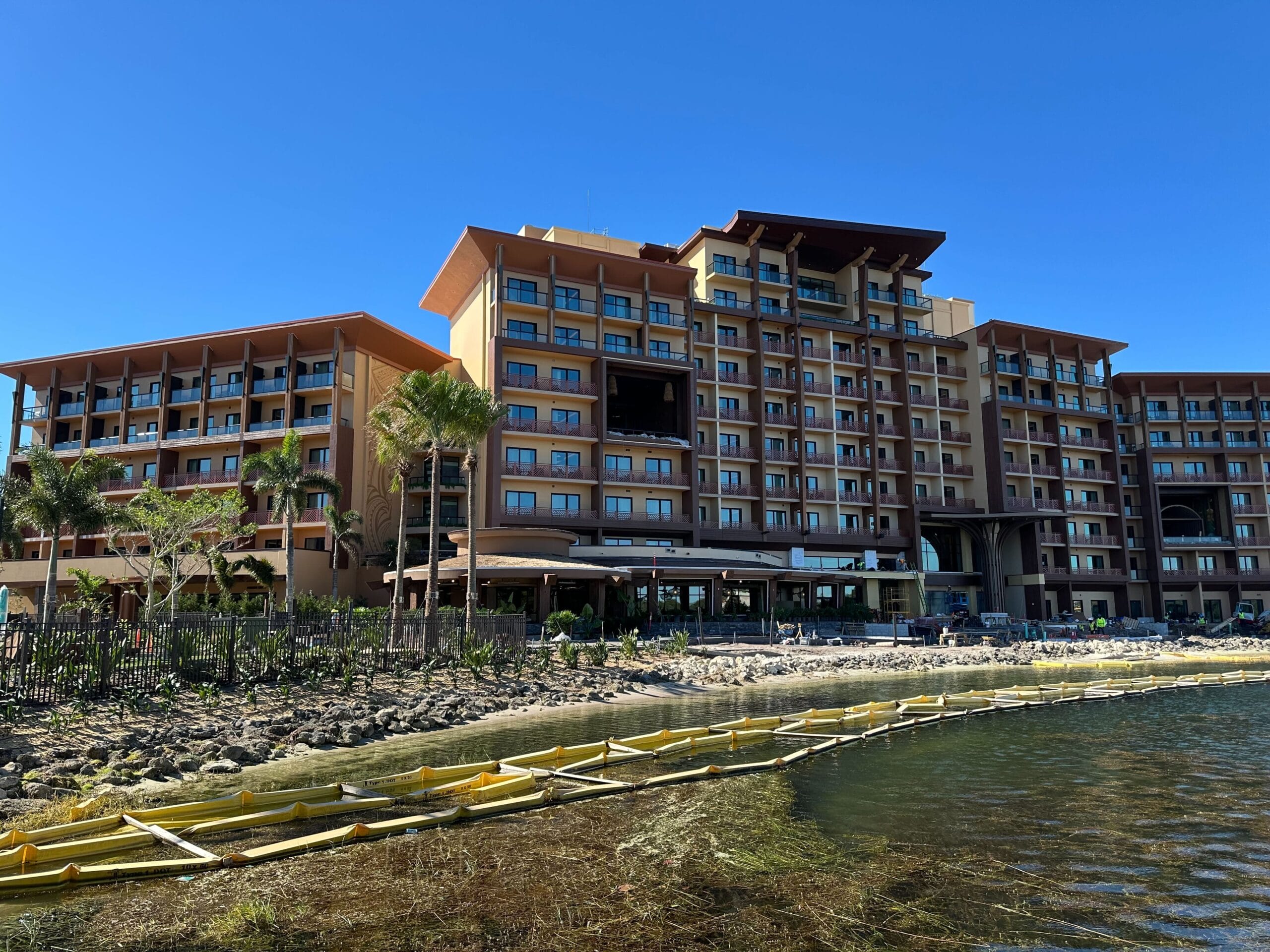 A multi-story hotel building reminiscent of Disney's Polynesian Village Resort offers a stunning waterfront view, surrounded by palm trees and adorned with vibrant murals under a clear blue sky. Relax in style with elegant patio furniture that complements this tropical paradise.