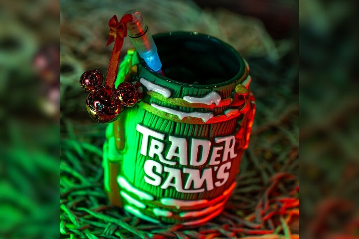 A green Trader Sam's Holiday Rum Barrel mug with a straw and decorative Mickey Mouse ornament is set on a textured surface, illuminated by festive red and green lights.