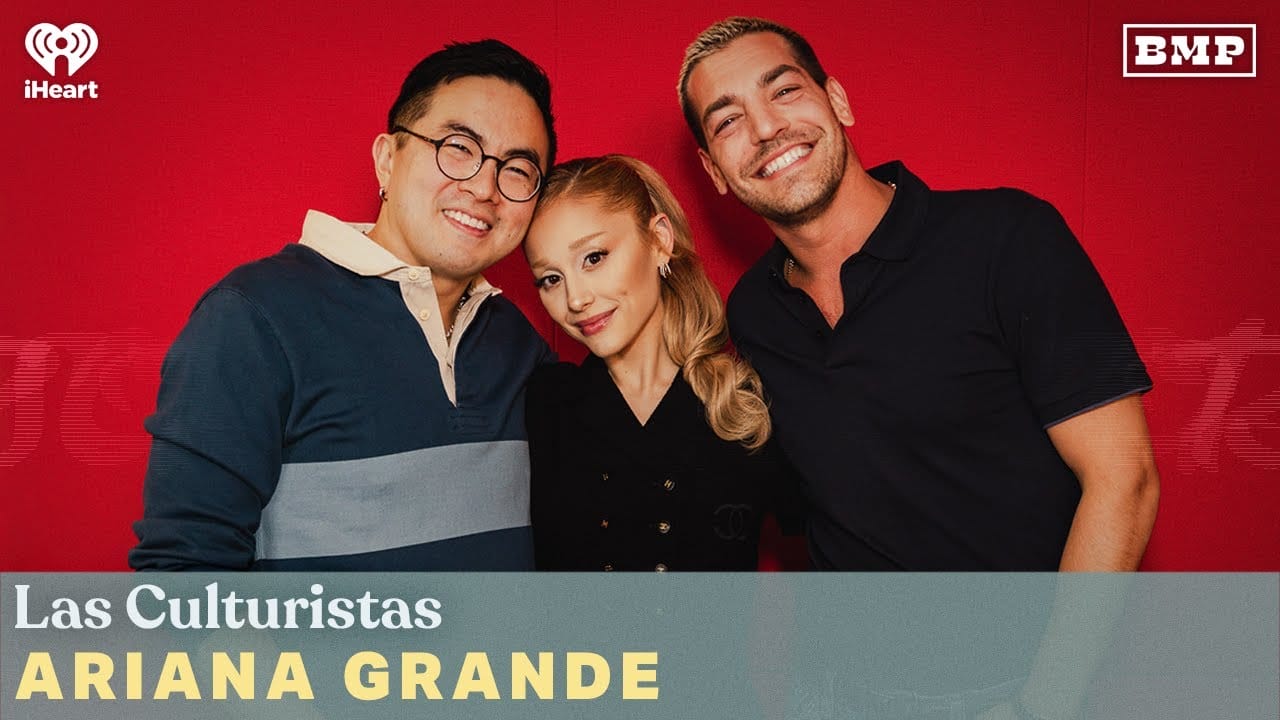Three people stand together in front of a red backdrop with the text "Las Culturistas" and "Ariana Grande.