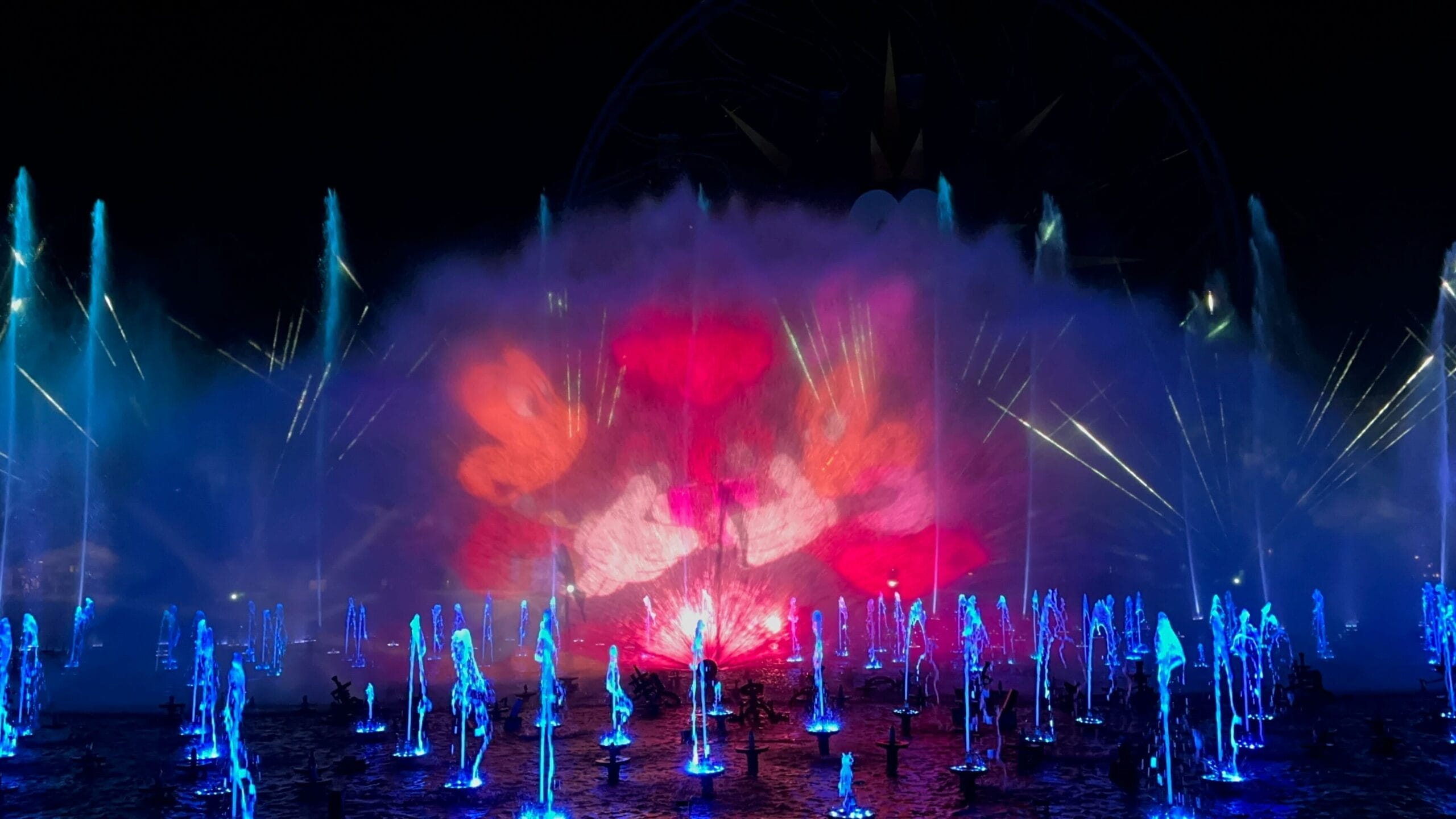 A scene from Mickey & Minnie's Gift of the Magi is projected onto a water screen at night, as water fountains illuminated blue bubble up from the ground.
