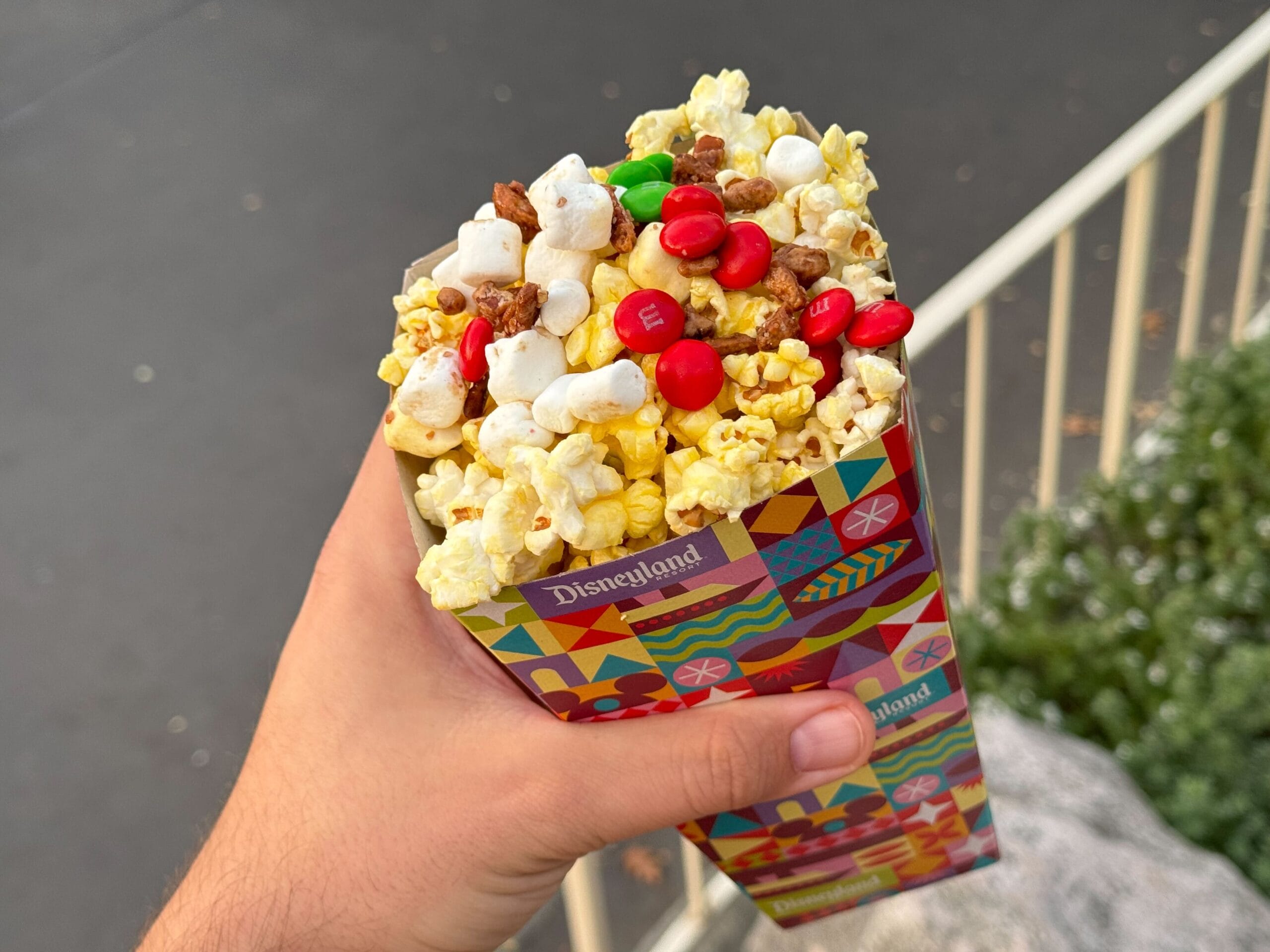 A person holds a Disneyland popcorn box filled with popcorn, M&M's, pretzels, and marshmallows, reminiscent of the sweet treats just steps away at Stage Door Cafe, all set against a blurred background.