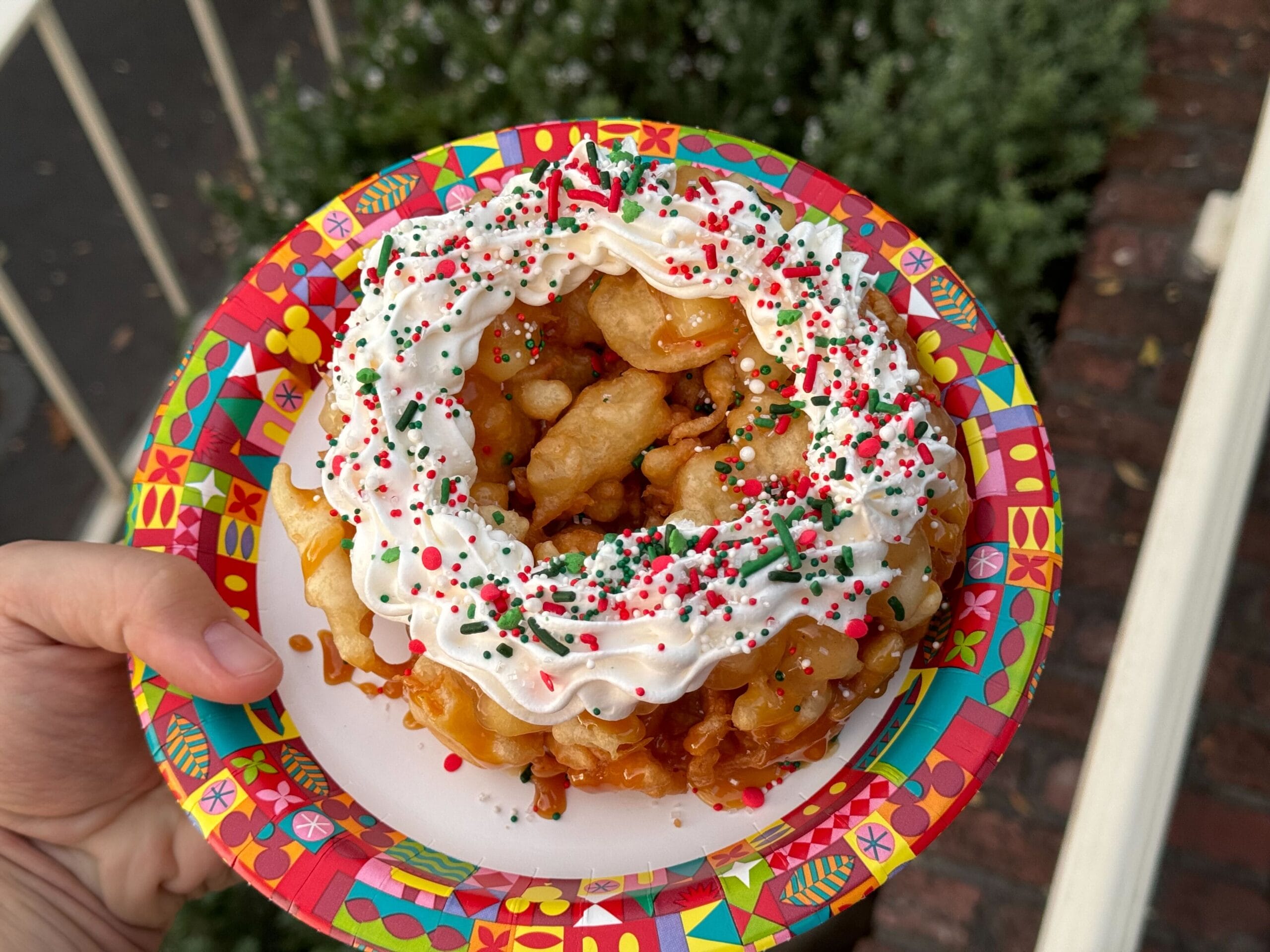A funnel cake topped with whipped cream and red, green, and white sprinkles is served on a colorful festive plate at the Stage Door Cafe.
