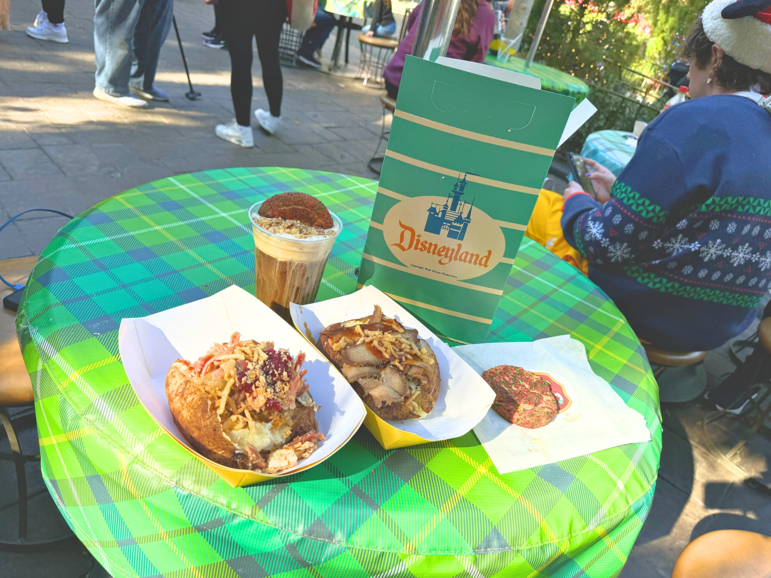 On a checkered tablecloth lies a Disneyland bag alongside iced coffee, two paper trays brimming with delicious food from Troubadour Tavern, and a cookie. In the background, people are seated and strolling by, enjoying the lively atmosphere.