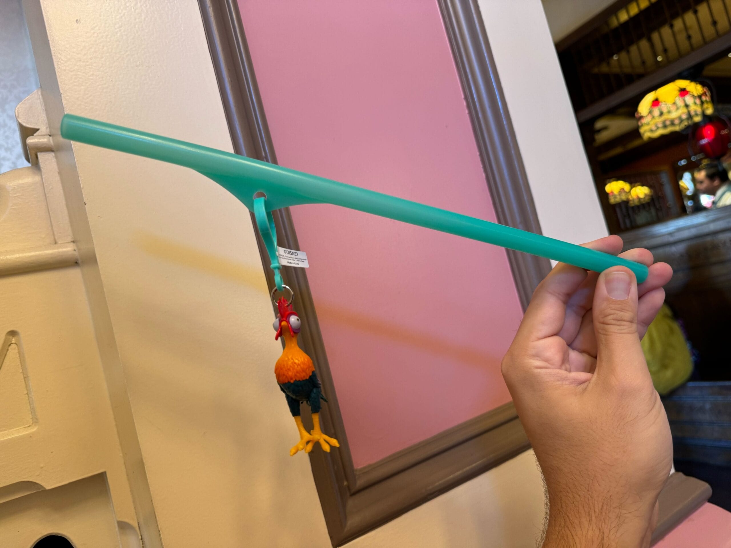 A hand grips a long green straw adorned with a Heihei straw clip, depicting a whimsical rooster figurine near the base. The background showcases walls with framed sections.