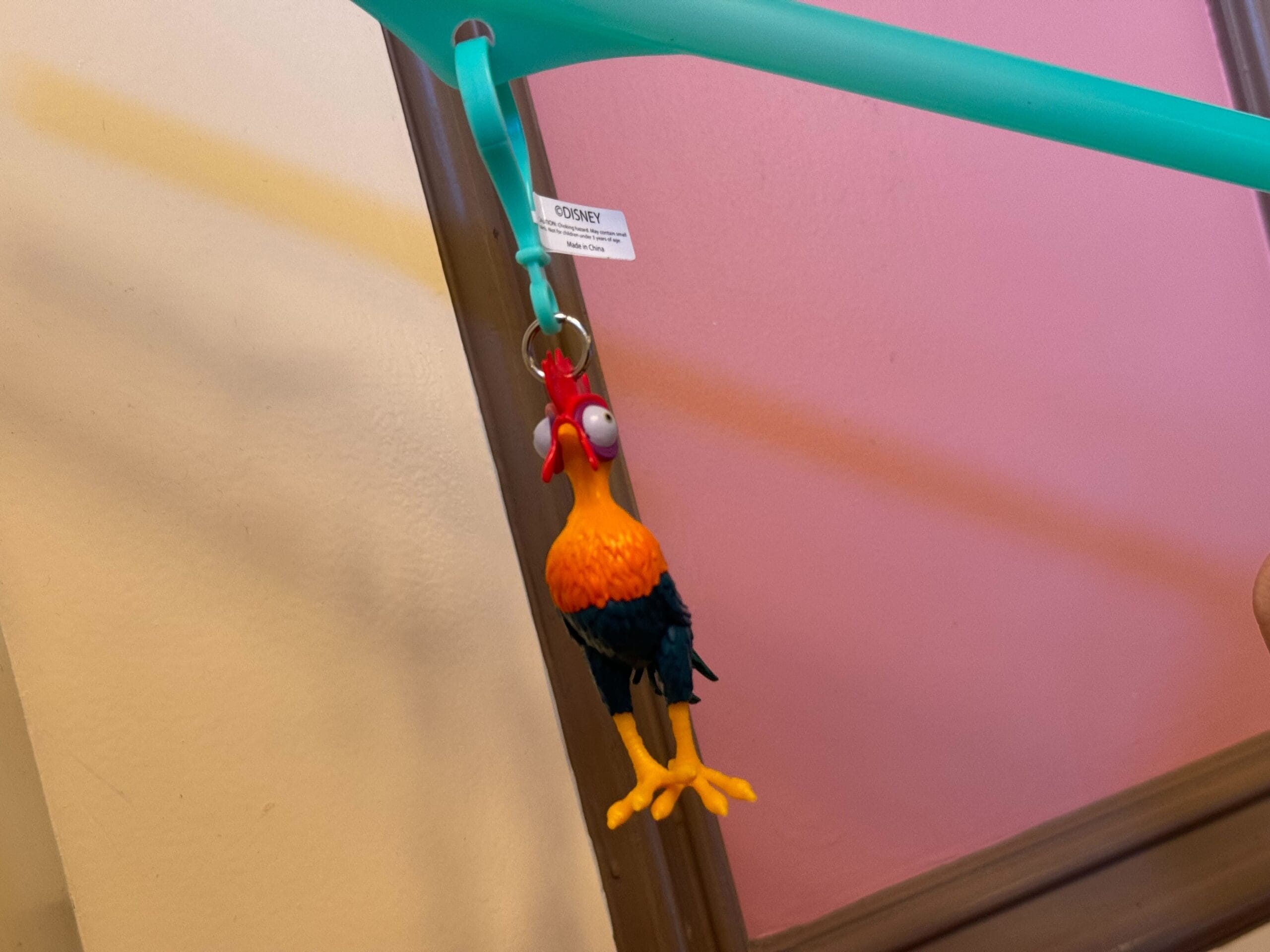 A colorful rubber chicken keychain and a cheeky Heihei straw clip dangle from a turquoise rod against a pink and beige wall.