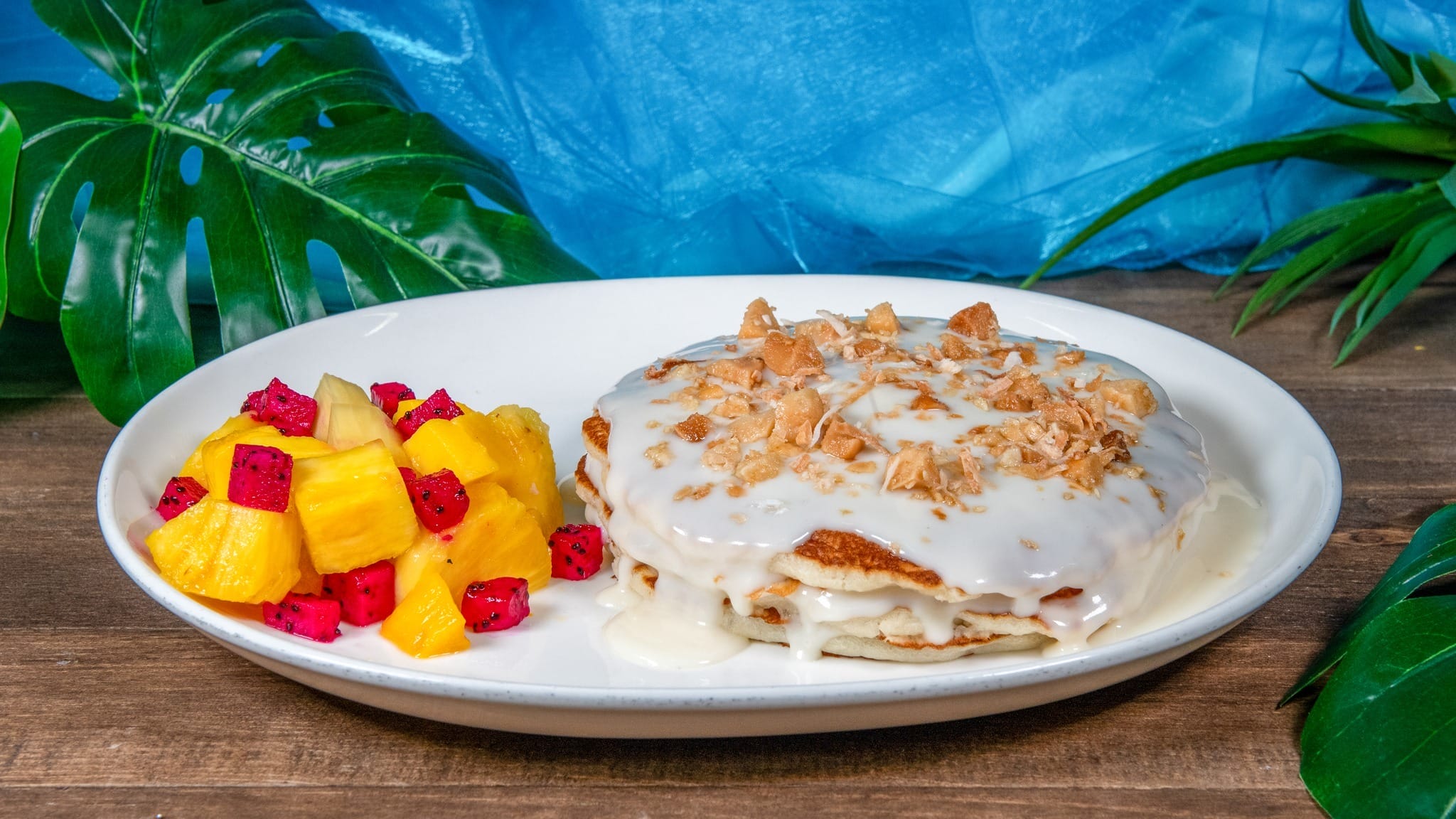 A plate of pancakes topped with coconut flakes and white sauce, served with fresh pineapple and dragon fruit cubes.