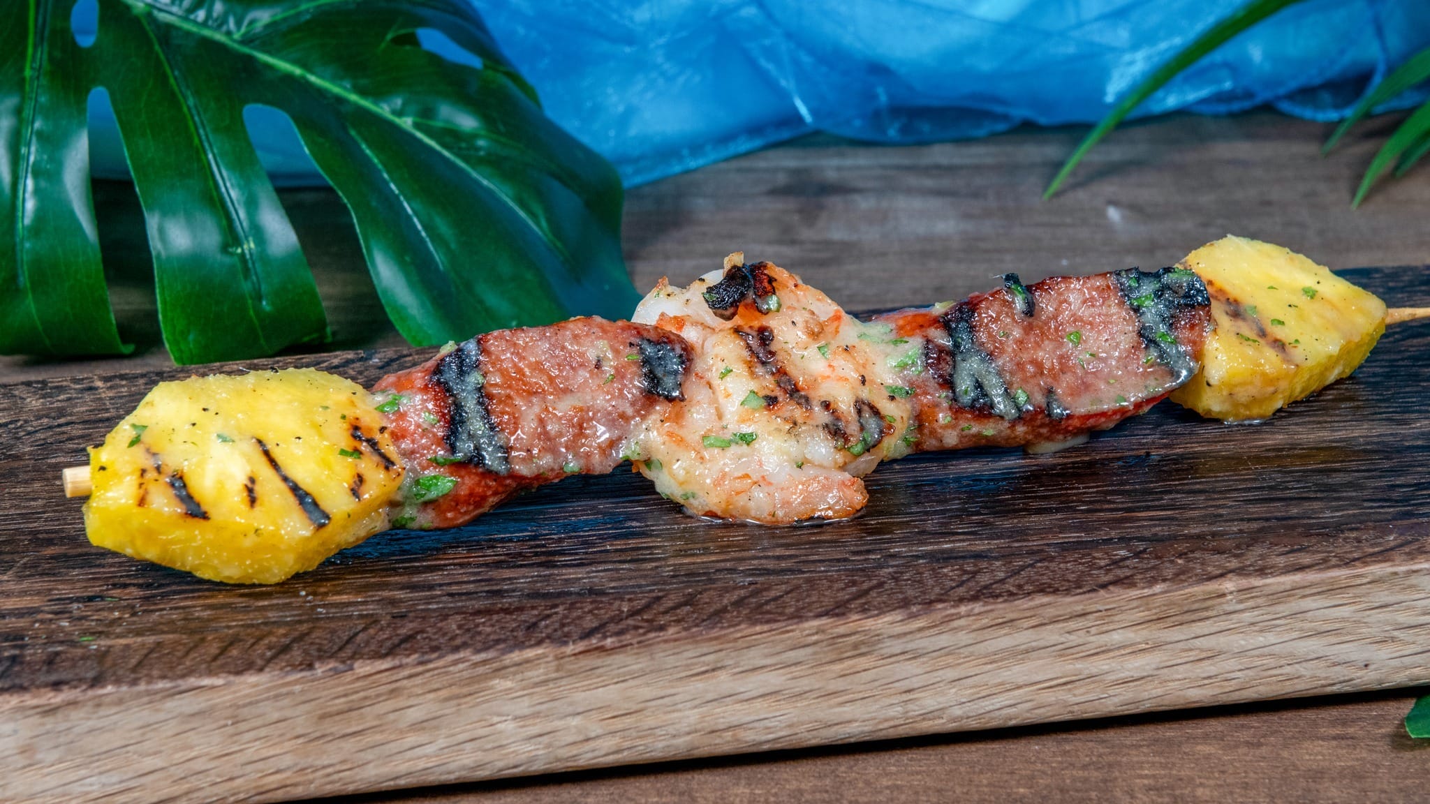 Grilled skewer with pineapple, shrimp, and meat on a wooden board, surrounded by green leaves.