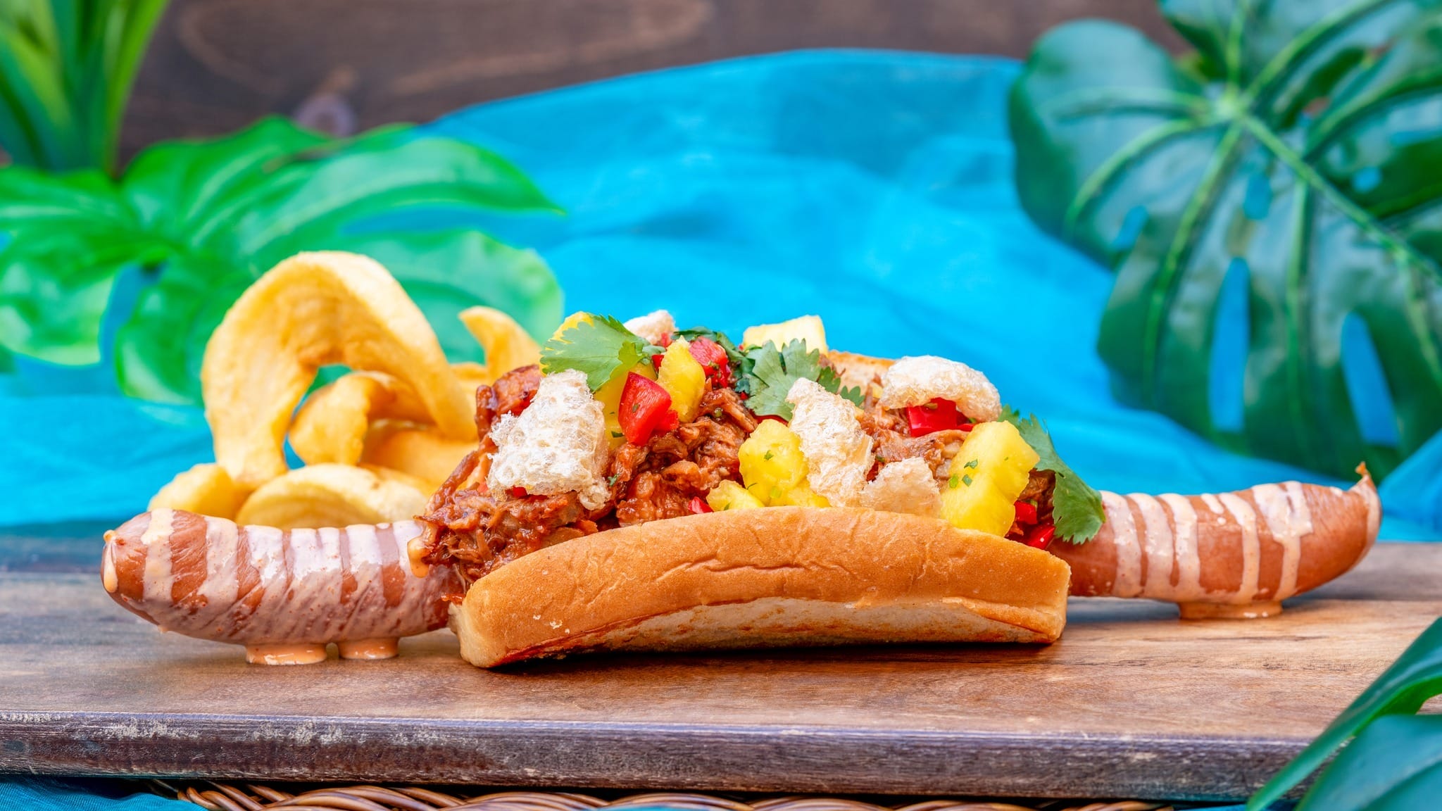 A hot dog with toppings and plantain chips on the side, set on a wooden board with green leaves and blue cloth in the background.