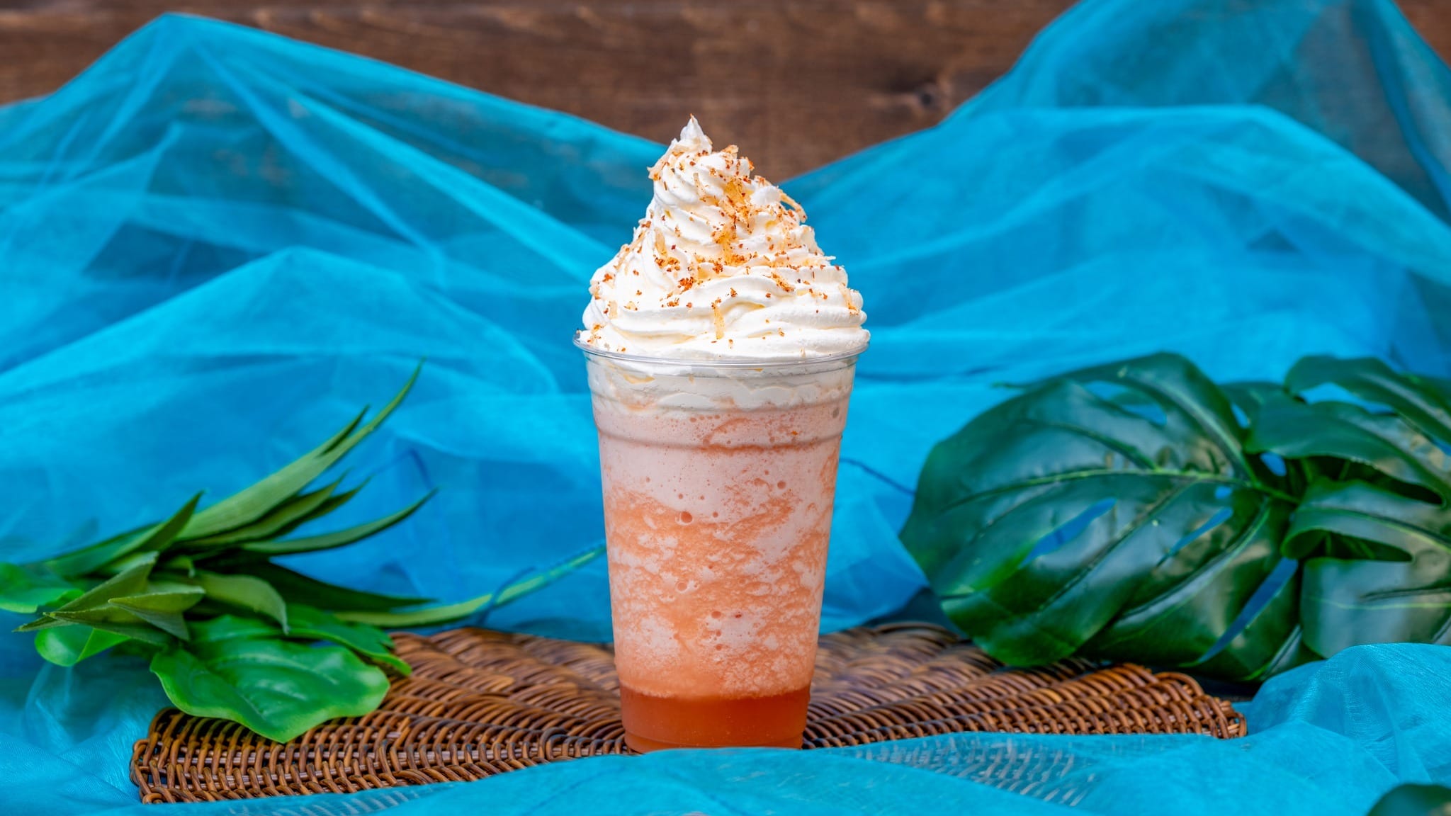 A tall glass of whipped cream-topped iced drink with orange and white layers, garnished with sprinkles, set against a blue background with green leaves.