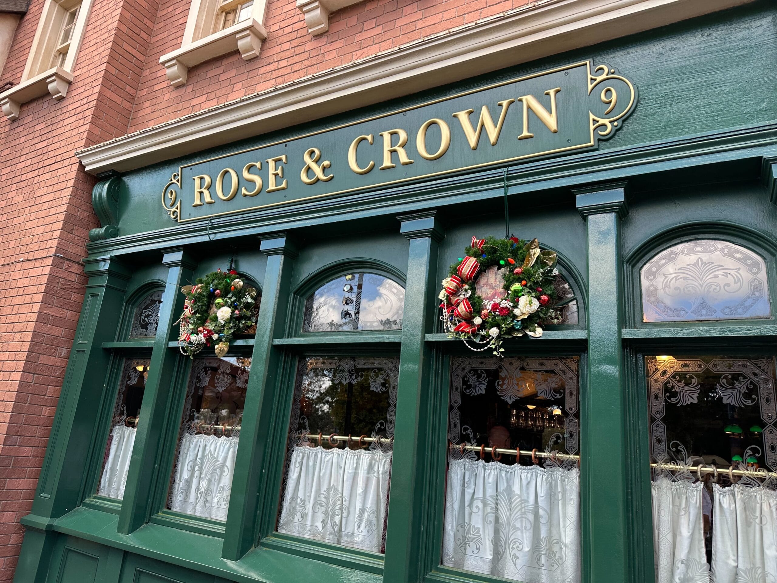 The exterior of the Rose & Crown pub captures a festive charm with its green facade, floral wreaths, and lace curtains, reminiscent of an Epcot Christmas tree's enchanting allure.