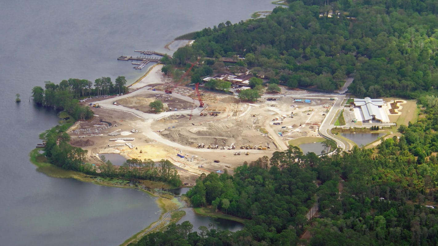 Aerial view of a construction site on a wooded peninsula by a lake, showcasing daily recap of progress with cleared land, equipment, and adjacent forested areas.