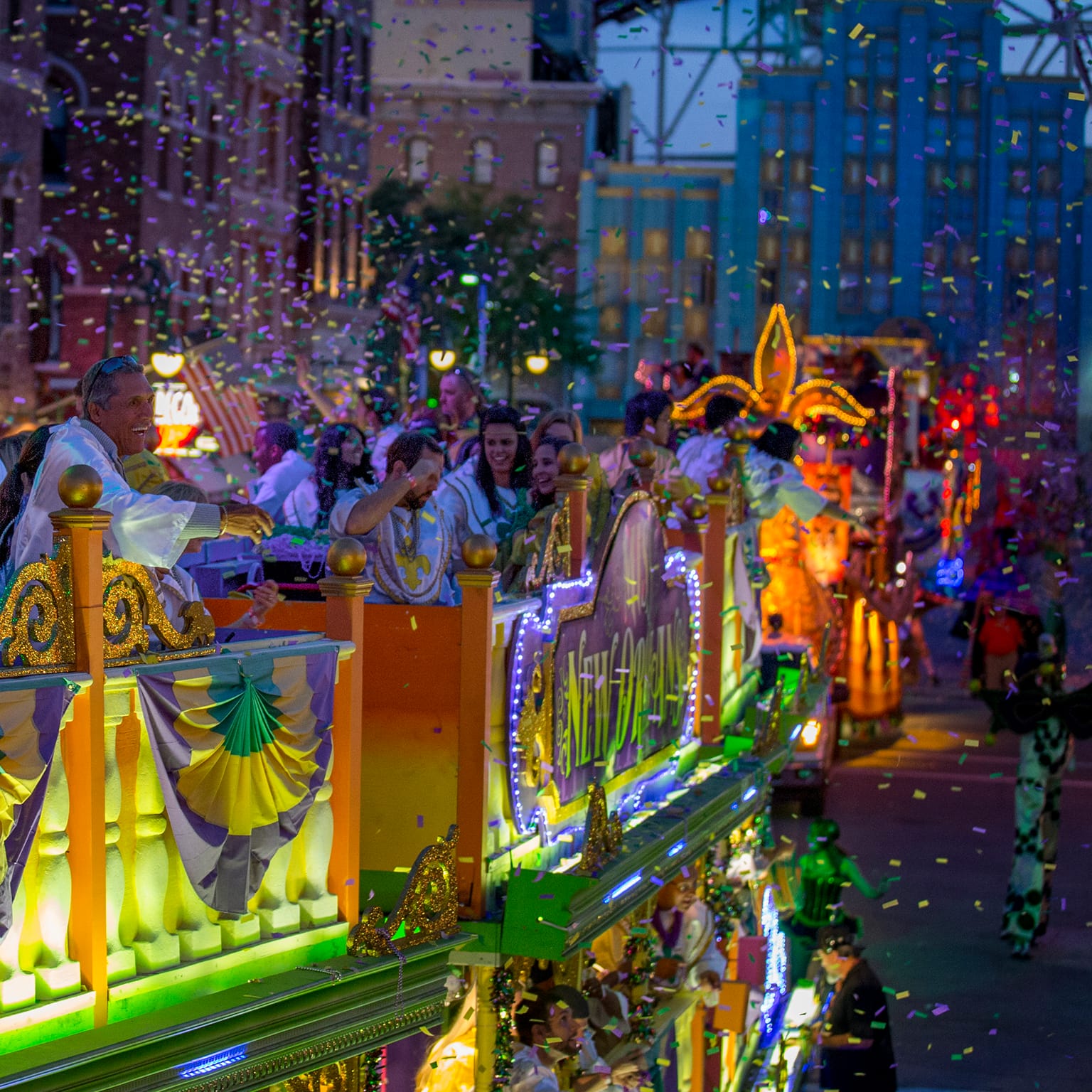 A colorful parade at night features a brightly lit float with people waving, surrounded by falling confetti and festive decorations.
