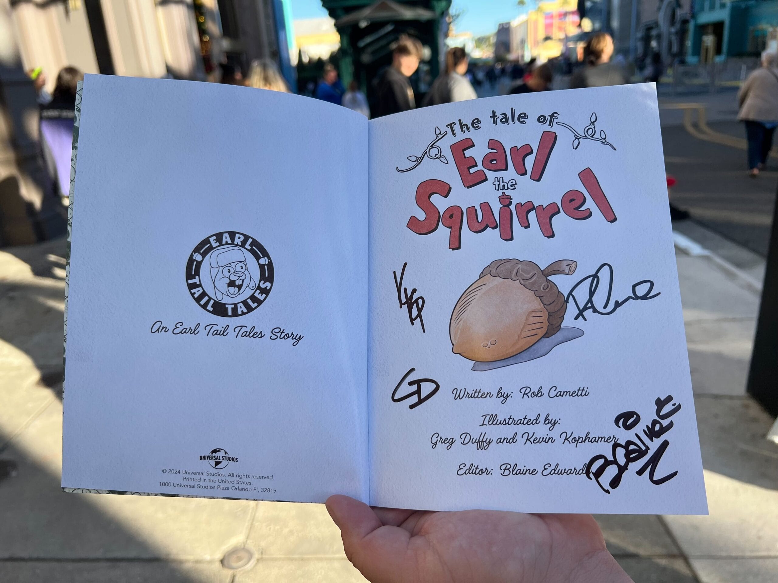 Two-page spread of a book titled "The Tale of Earl the Squirrel" with signatures and illustrations of an acorn and squirrel tail on the right page, held by a hand outside in a bustling street.