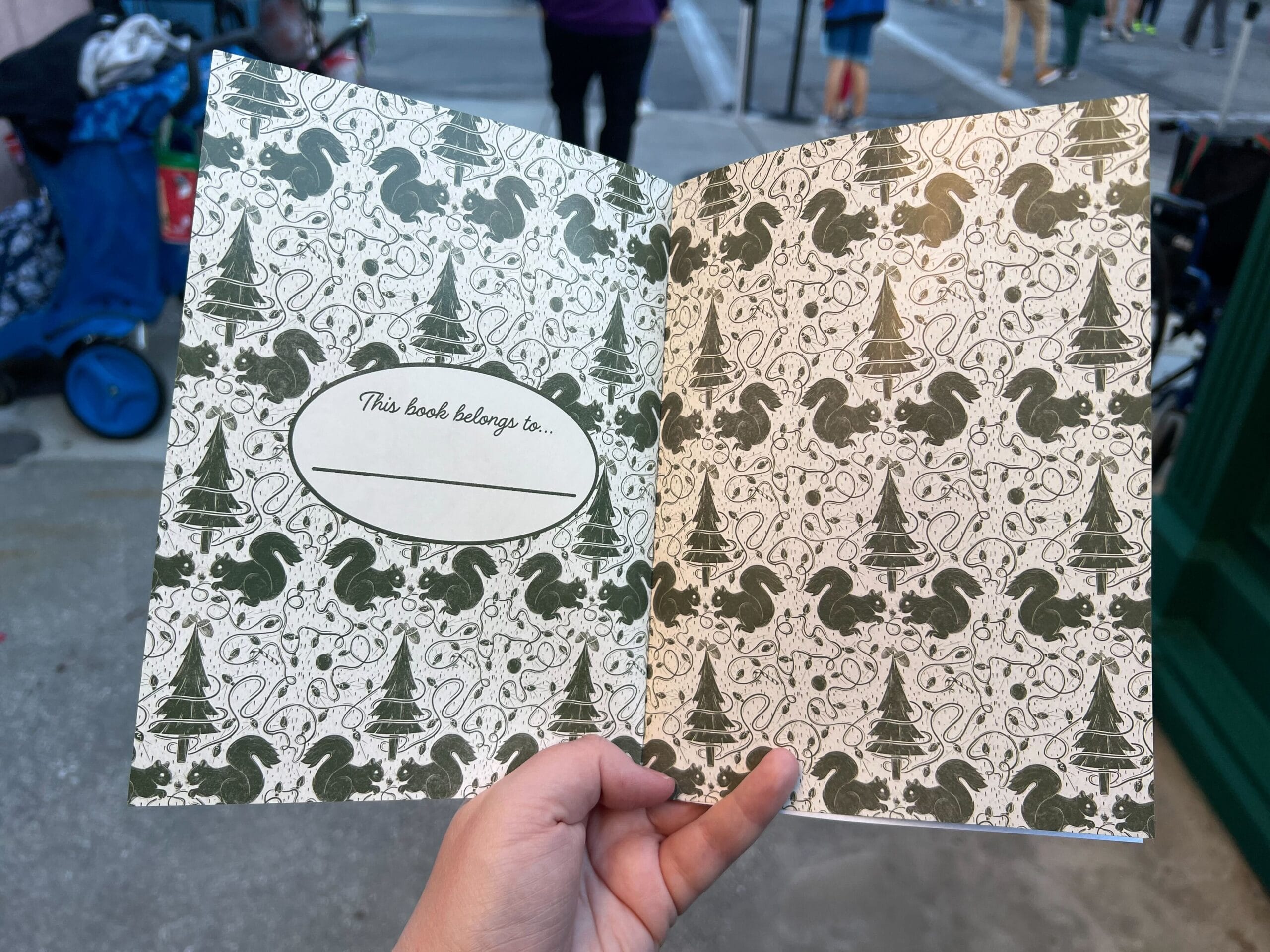Hand holding an open book with a decorative forest-themed pattern and a label that reads "This book belongs to..." in a city street setting.