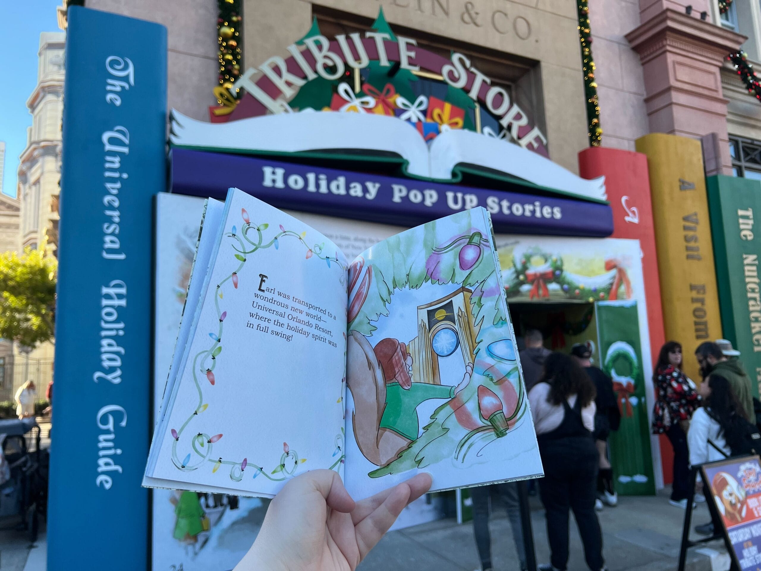A person holds an open illustrated book in front of a holiday-themed store with large decorative book spines and people entering and exiting the building.
