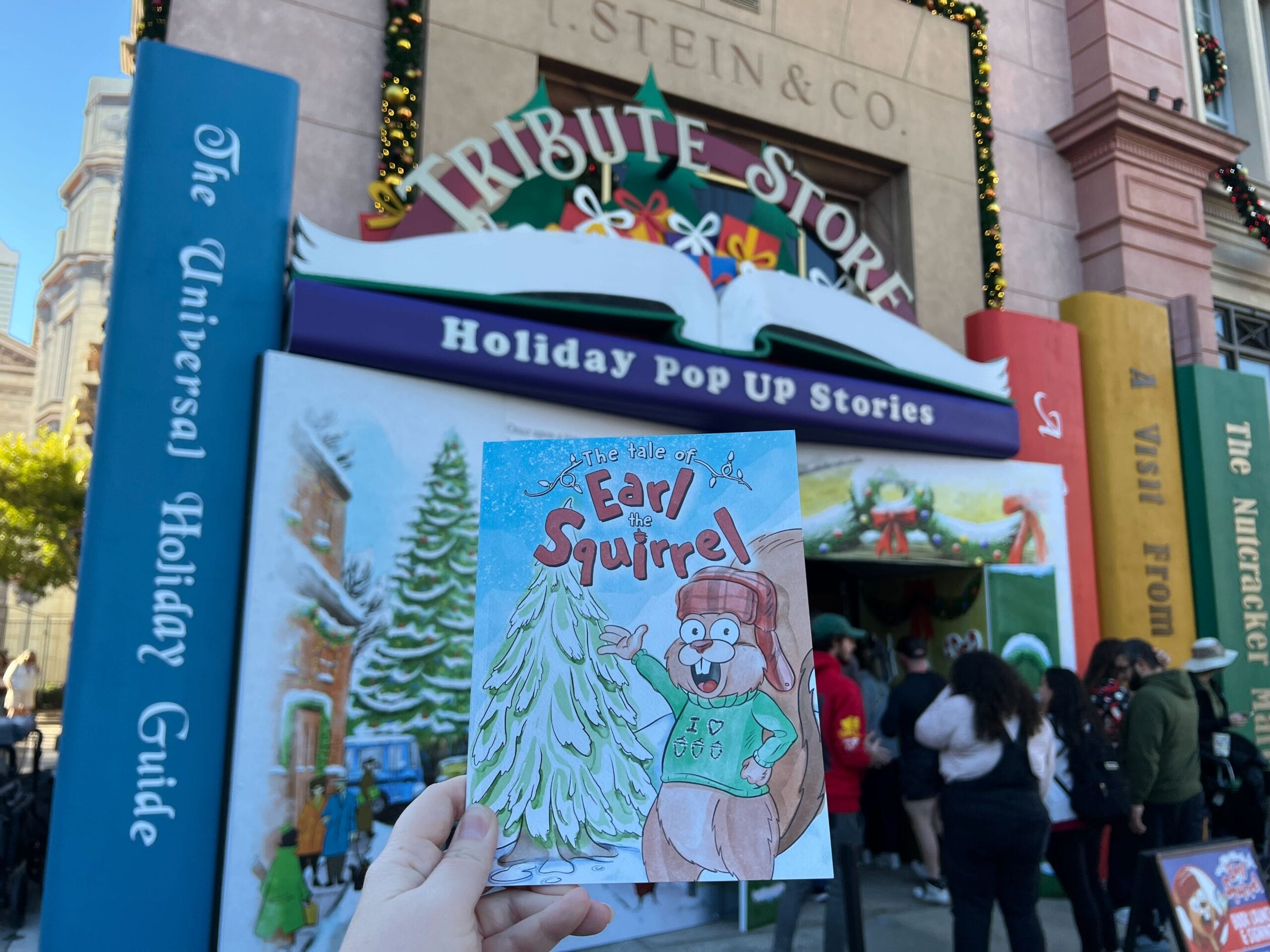 Hand holds "The Tale of Earl the Squirrel" book in front of a holiday-themed pop-up store entrance with people gathered outside.