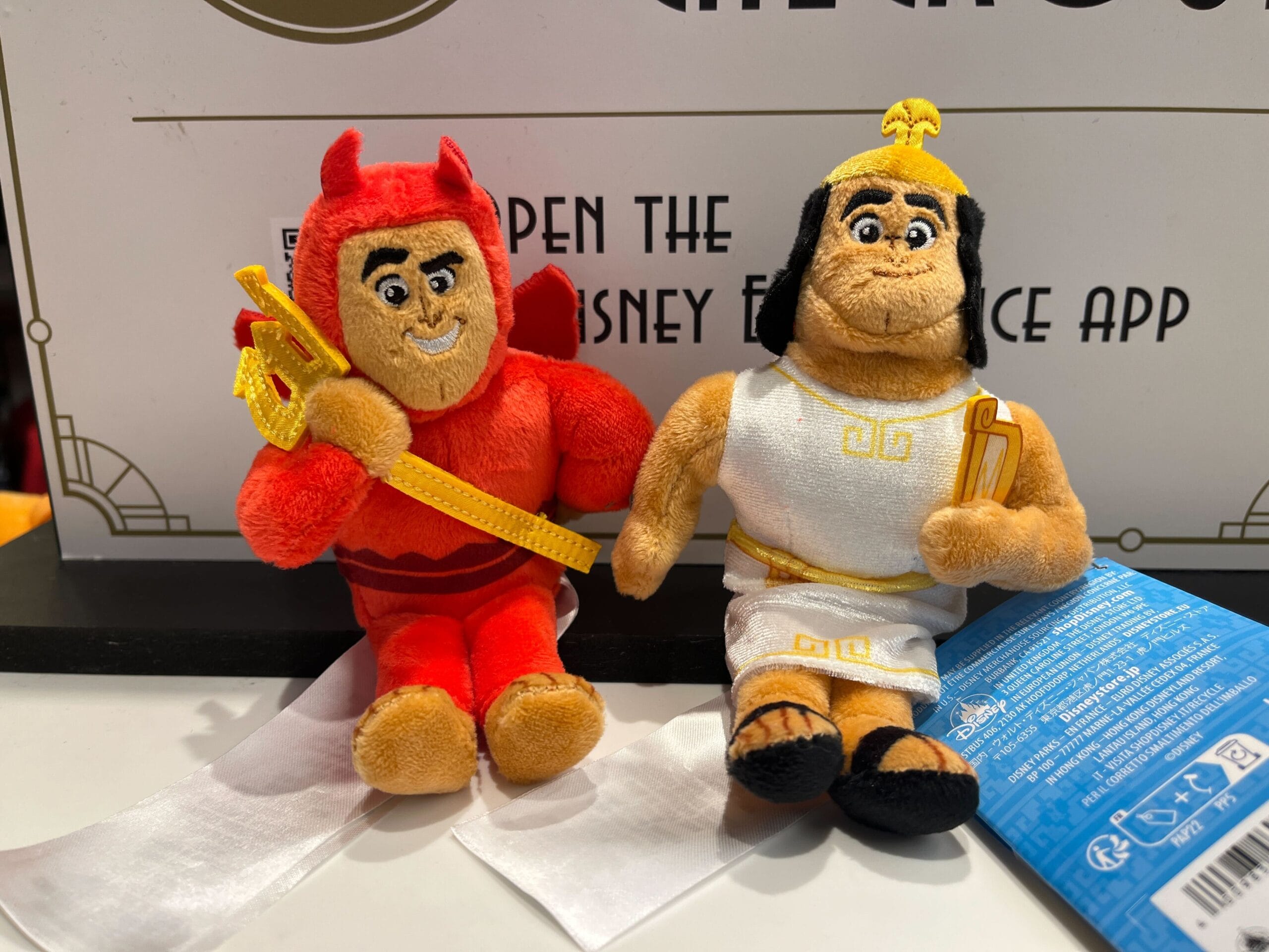 Two plush toys on display: one dressed as a red devil holding a trident, and the other as a Greek god in a white toga holding a lightning bolt.