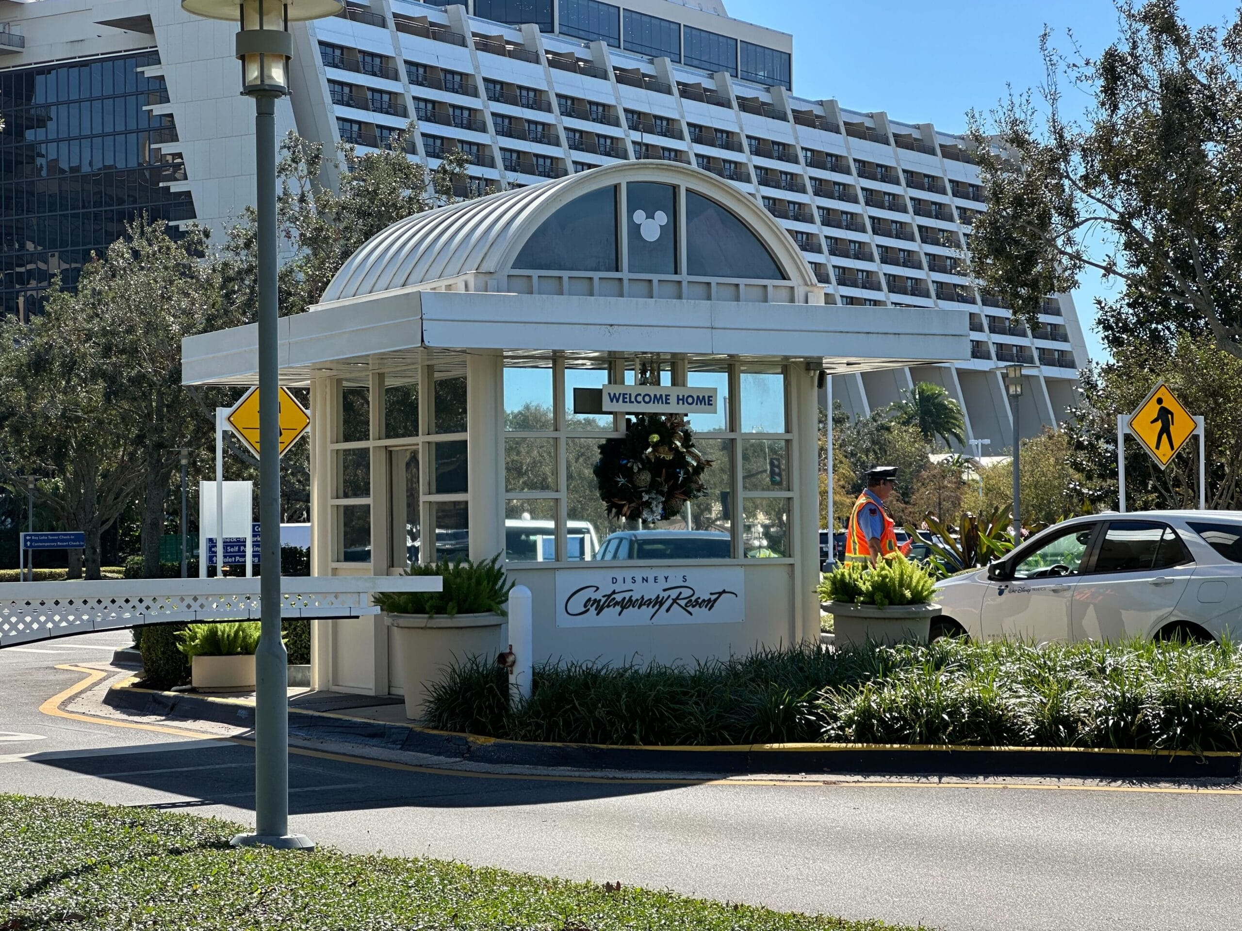 A security booth at a resort entrance with a "Welcome Home" sign and Disney's Contemporary Resort logo. A security guard stands nearby as a car approaches.