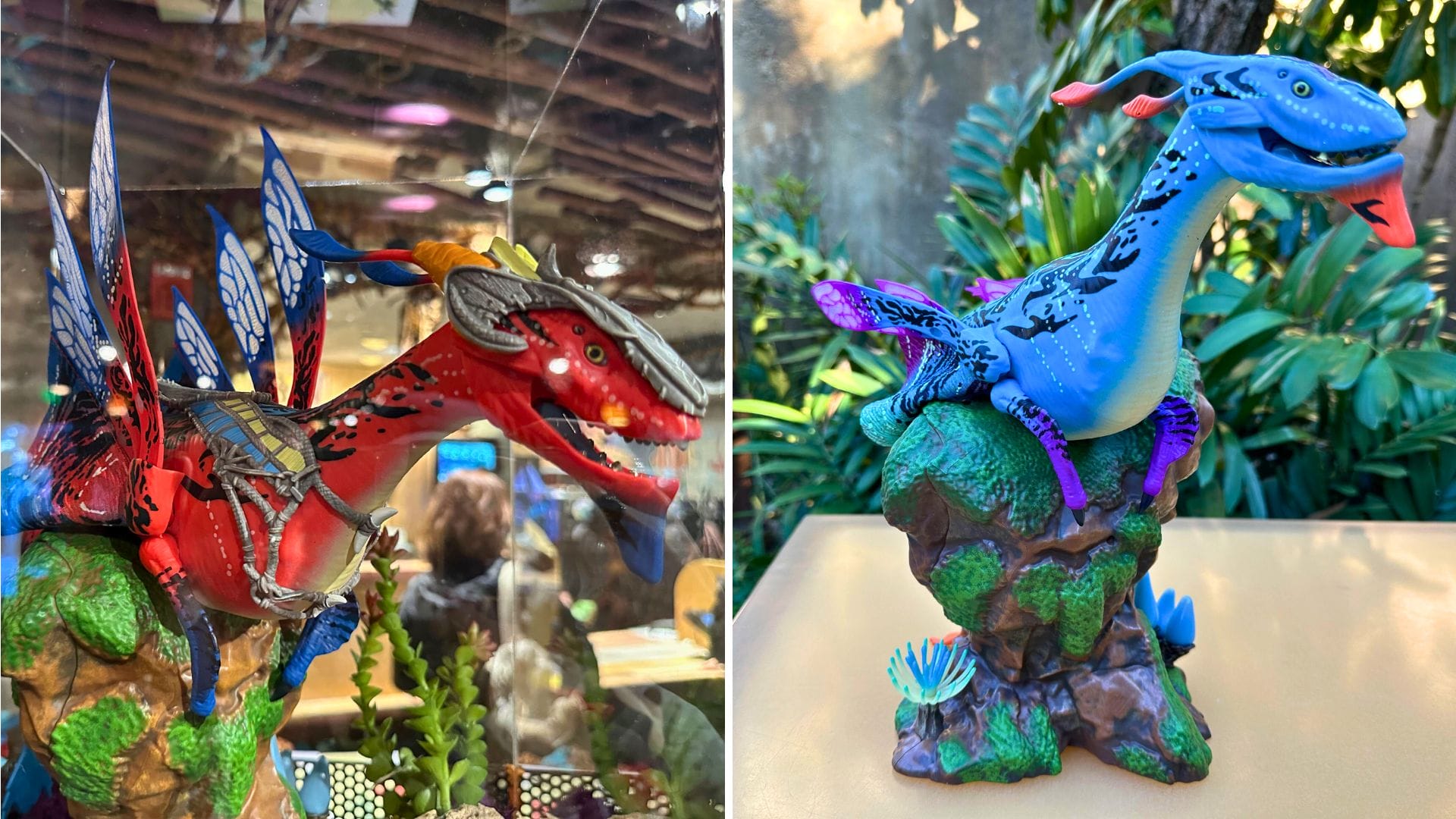 Two colorful dragon figurines are displayed. The left dragon is red with blue wings and the right one is blue with purple details, both set on rocky bases amidst plants.