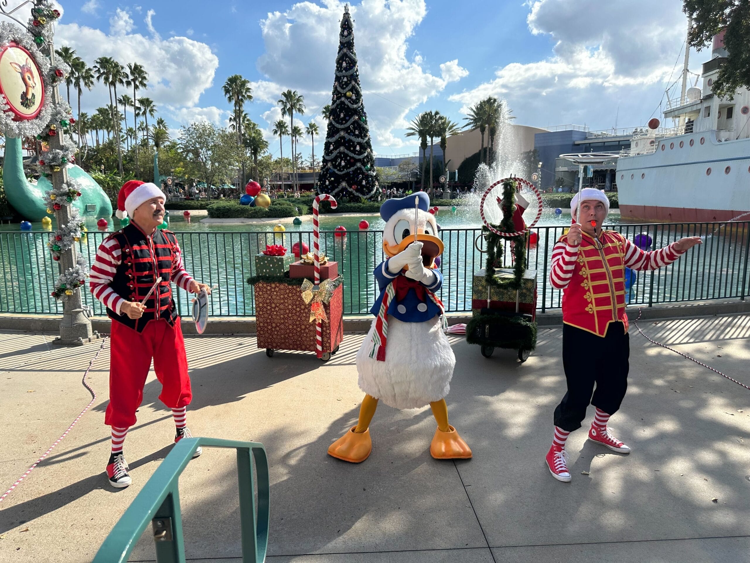 Donald Duck and two performers in festive clothing entertain guests at a Christmas party beside a decorated tree near a water feature on a sunny day, capturing the essence of holiday entertainment at Disney's Hollywood Studios.