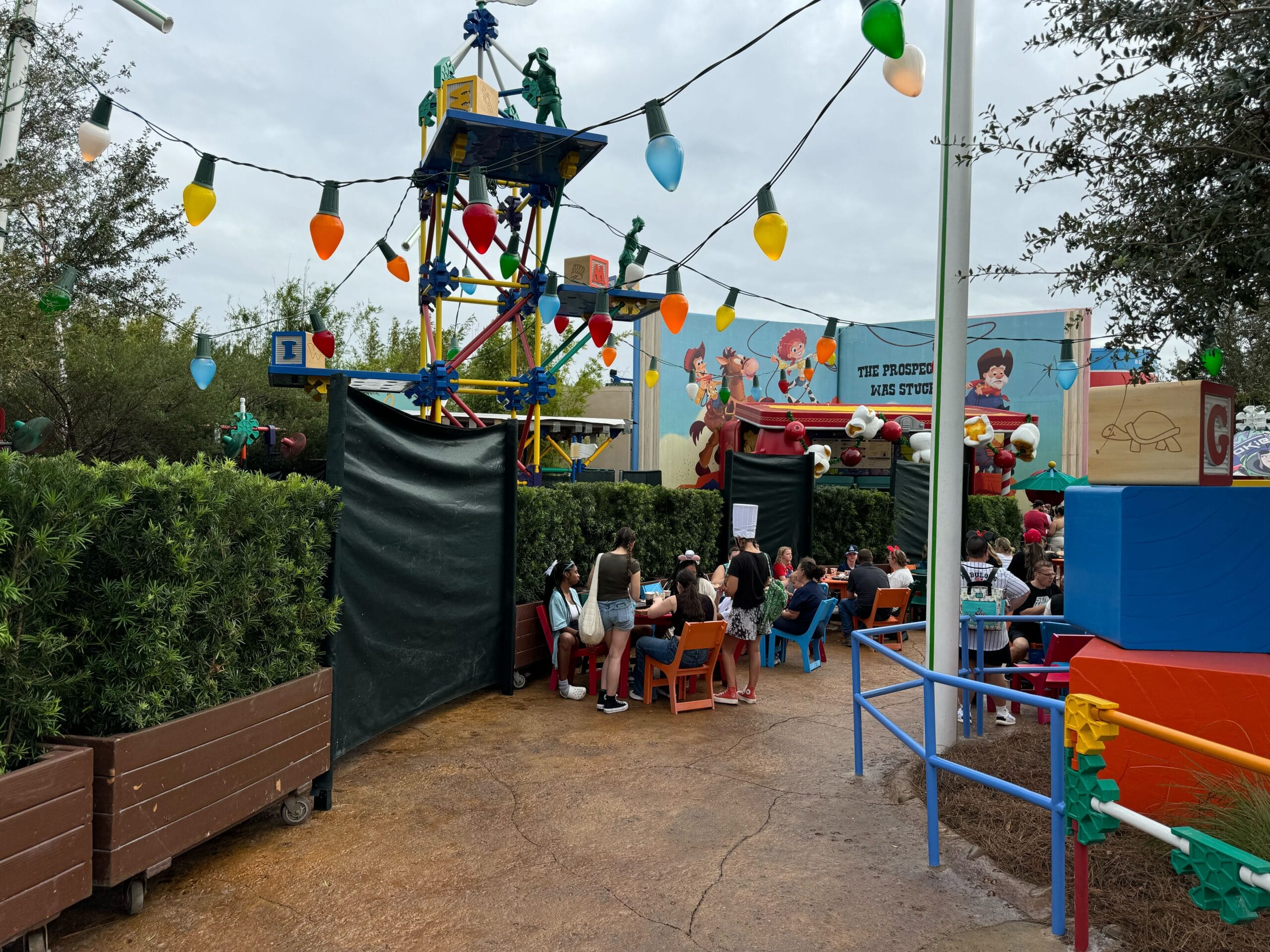 People sit at outdoor tables under string lights near colorful toy-themed decorations and green foliage.