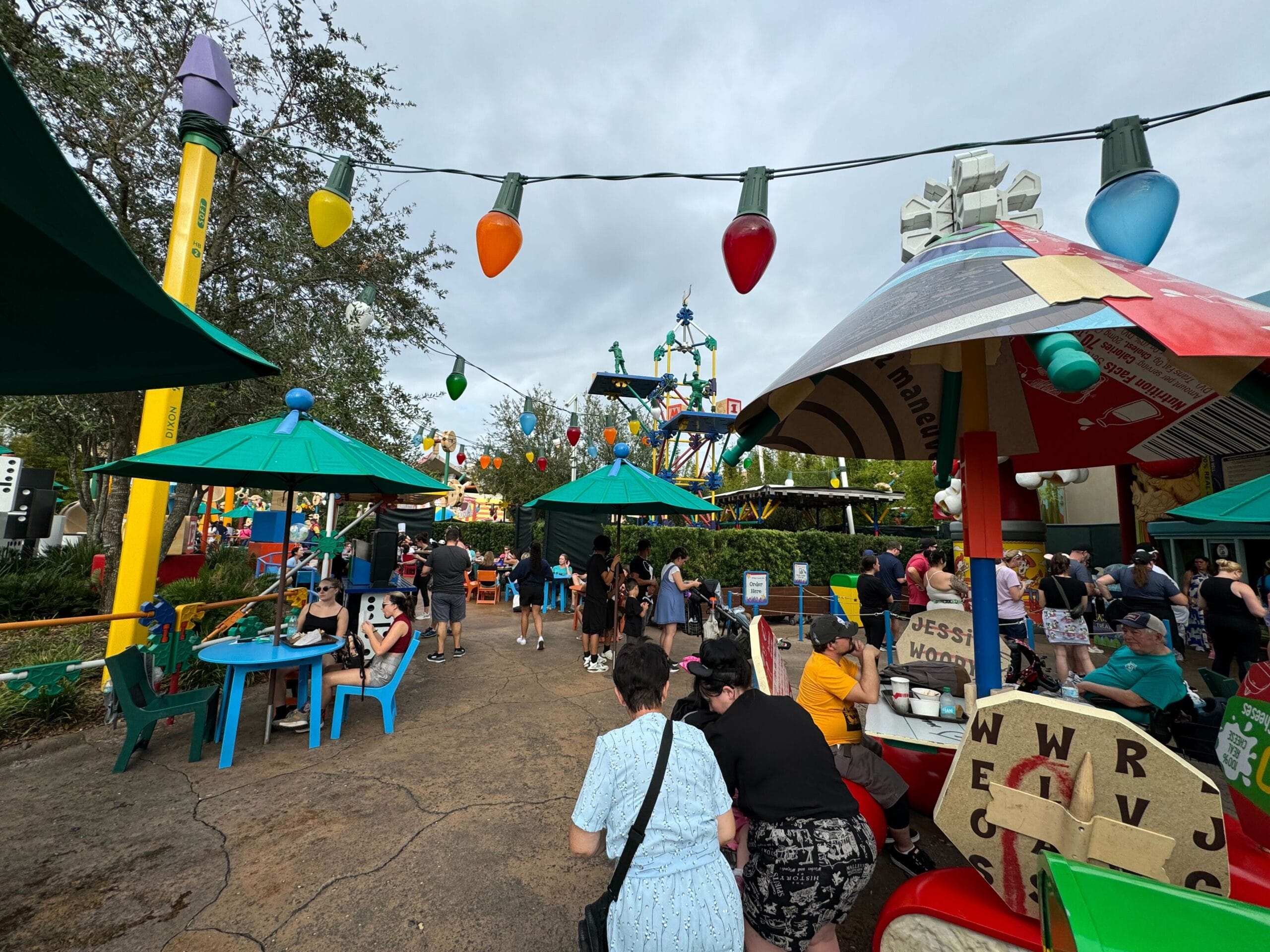 People walking and sitting in a vibrant amusement park area with colorful decorations and a game booth nearby.