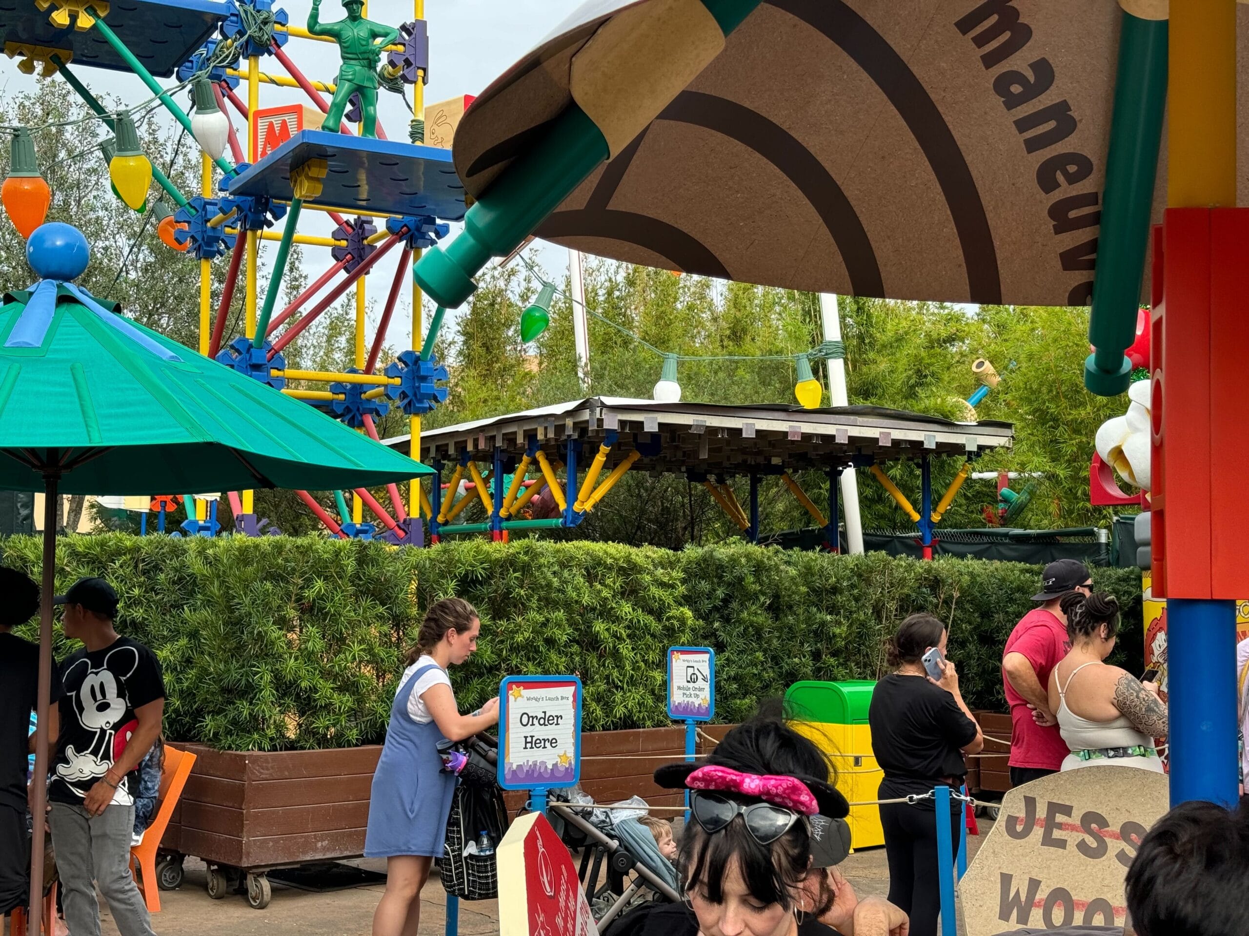 People waiting in line at an outdoor theme park attraction, surrounded by colorful umbrellas and play structures, with greenery in the background.