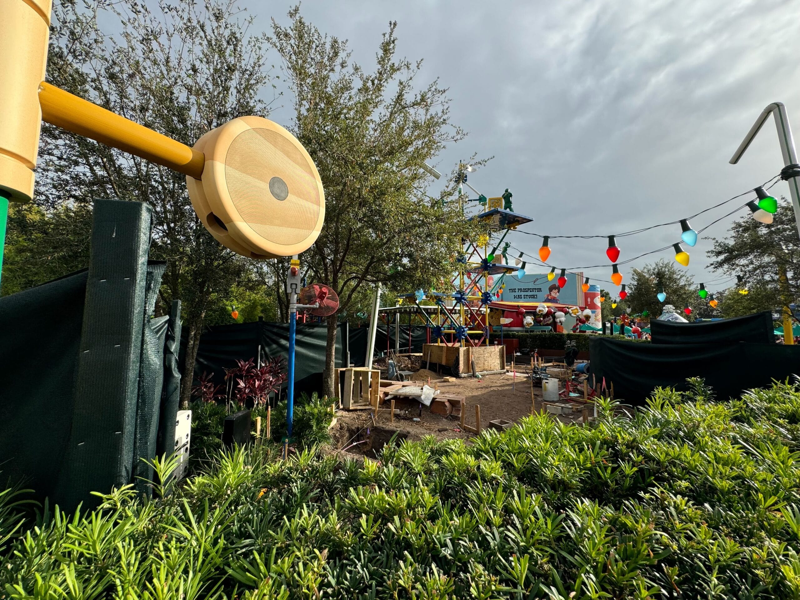Amusement park under construction with colorful structures, lights, and greenery in the foreground. Cloudy sky overhead.