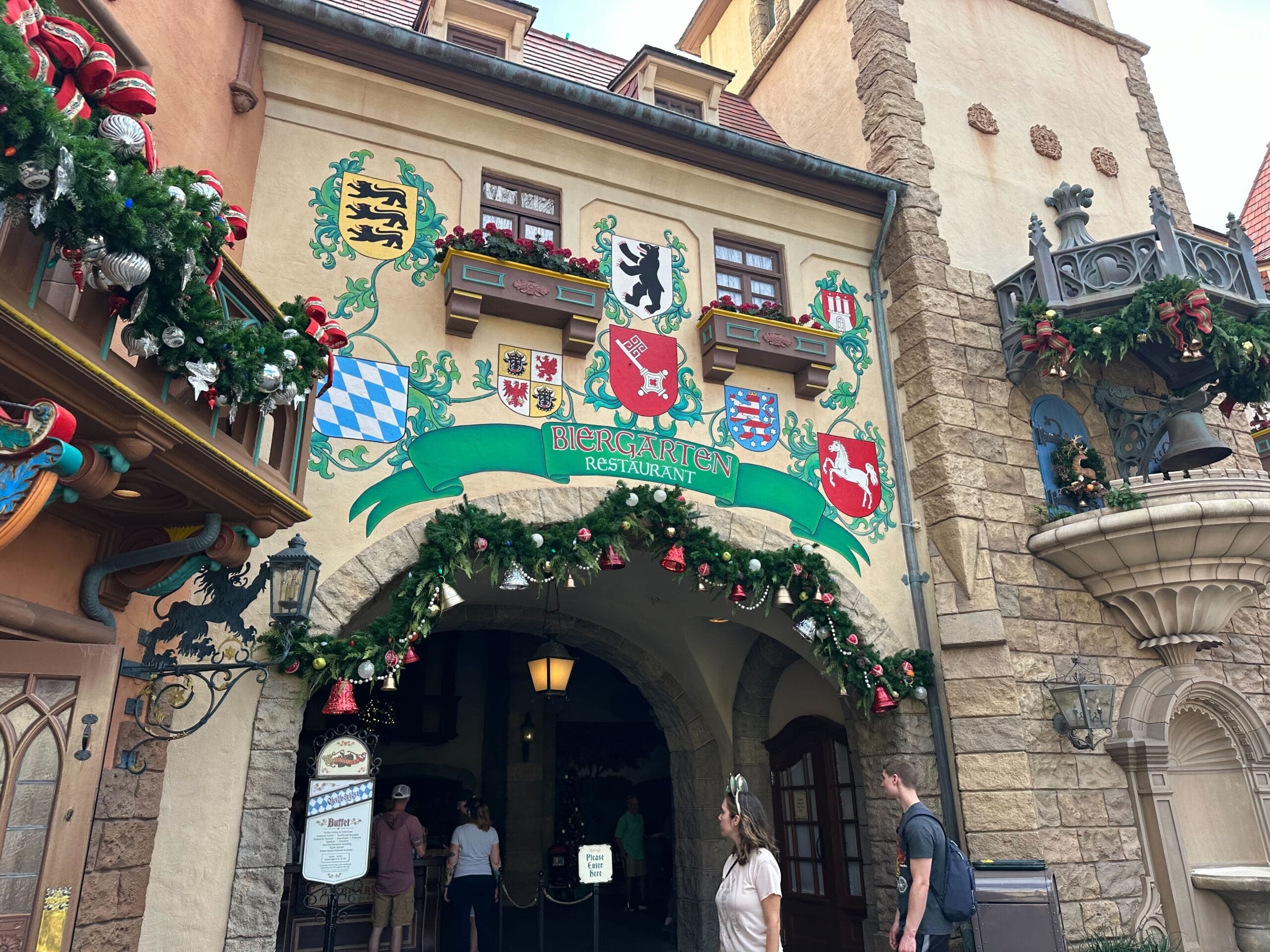 Entrance to Biergarten Restaurant, decorated with festive garlands and various coat of arms, featuring a stone archway and a menu board on the left. Nearby, Epcot's Christmas tree adds a touch of holiday magic to the scene.