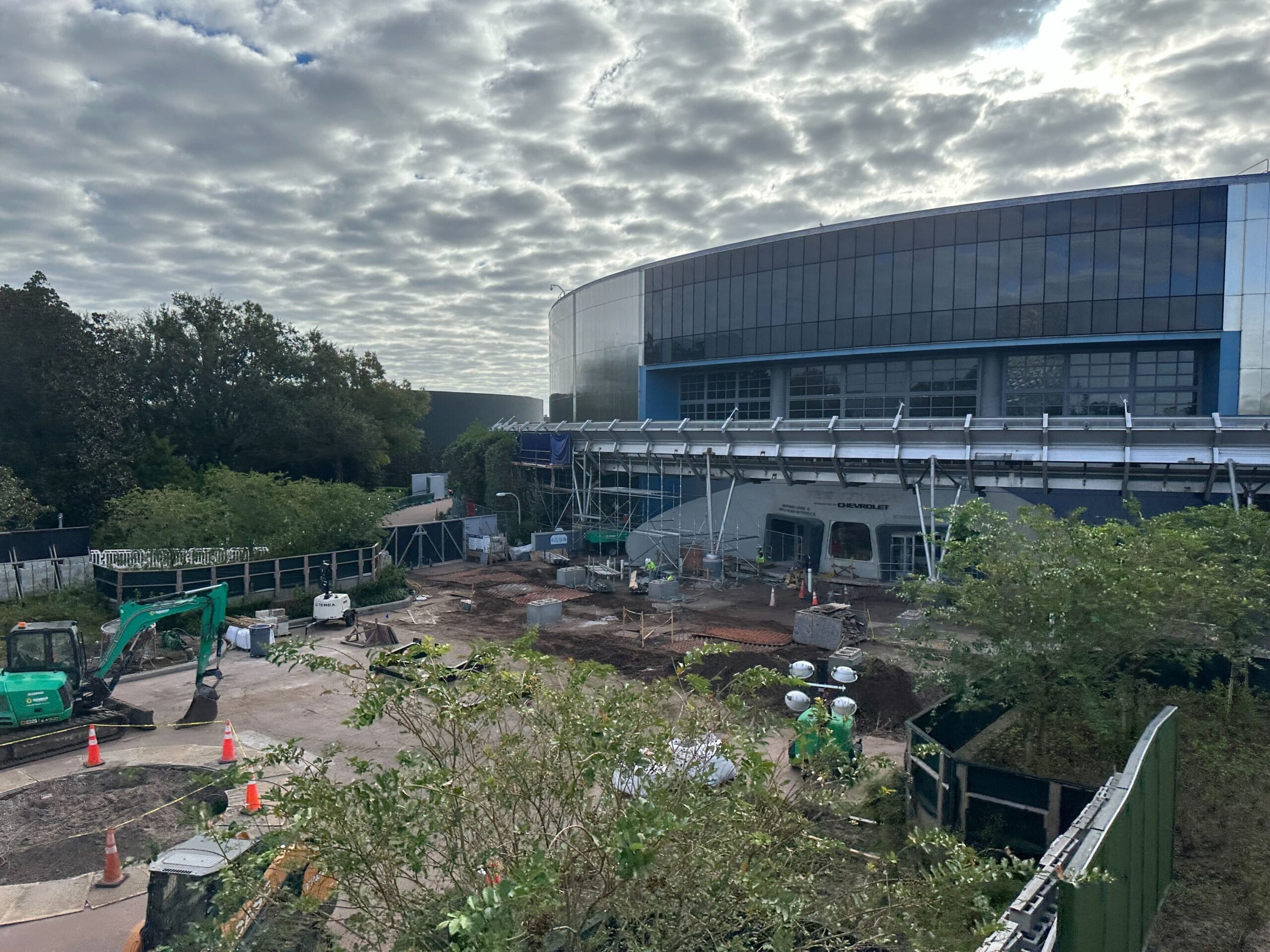 Construction site in front of a large, modern building with glass facade. Excavators and building materials scattered around. Overcast sky in the background.