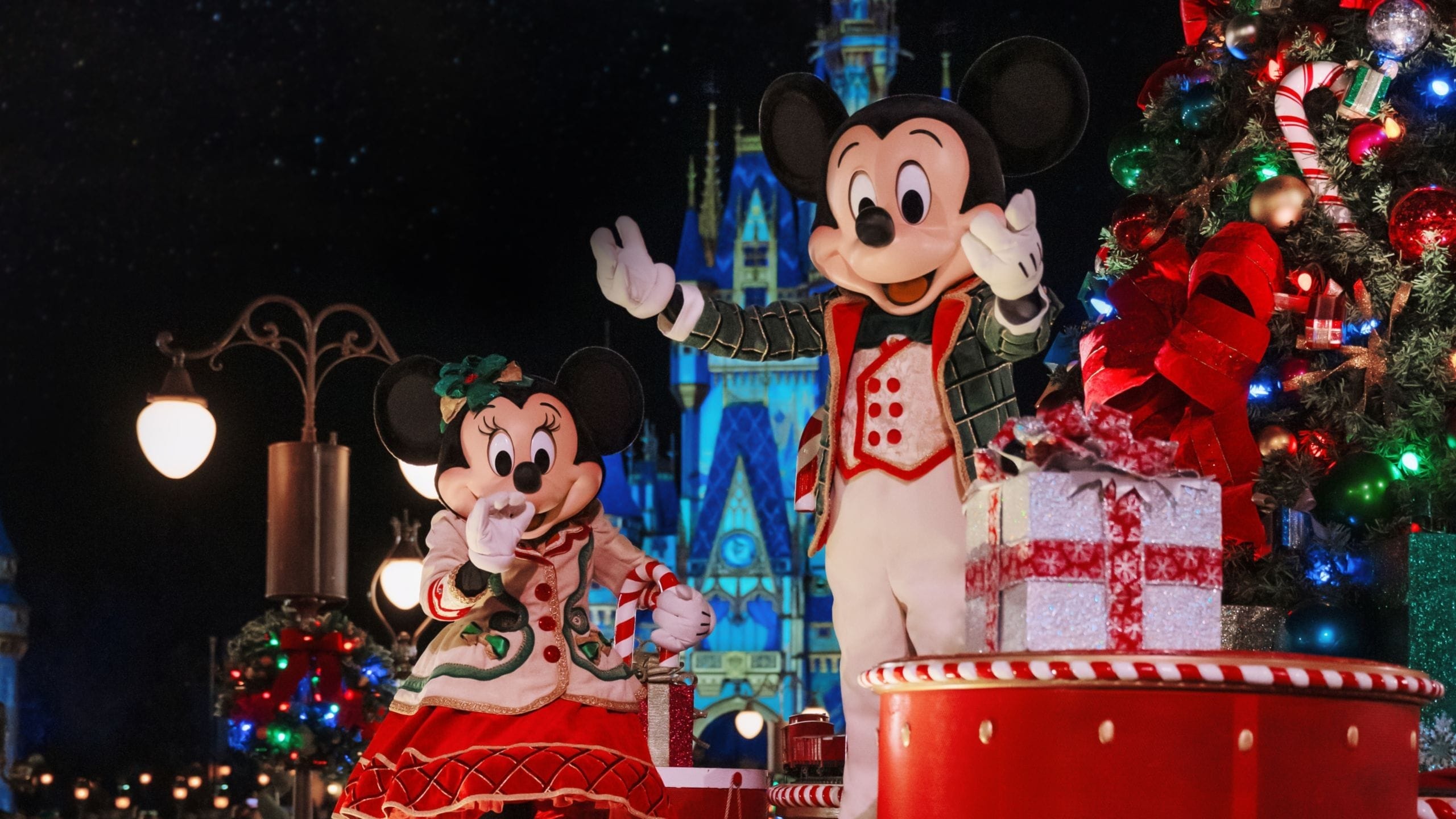 Mickey and Minnie Mouse in festive outfits stand beside a decorated Christmas tree, with a castle in the background at night.