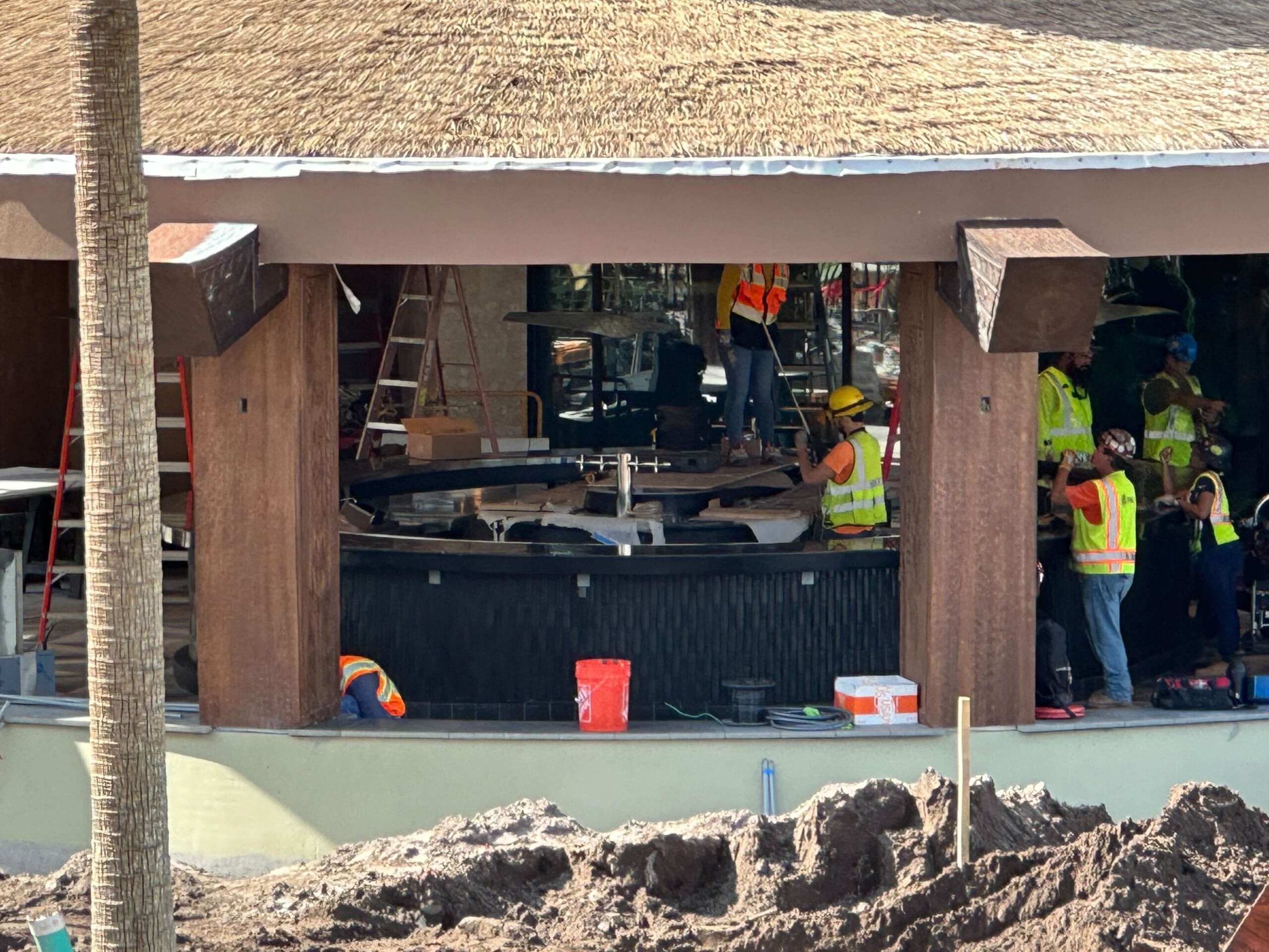 Construction workers in safety gear install a large circular structure under a thatched roof. Tools and equipment are scattered around the site.