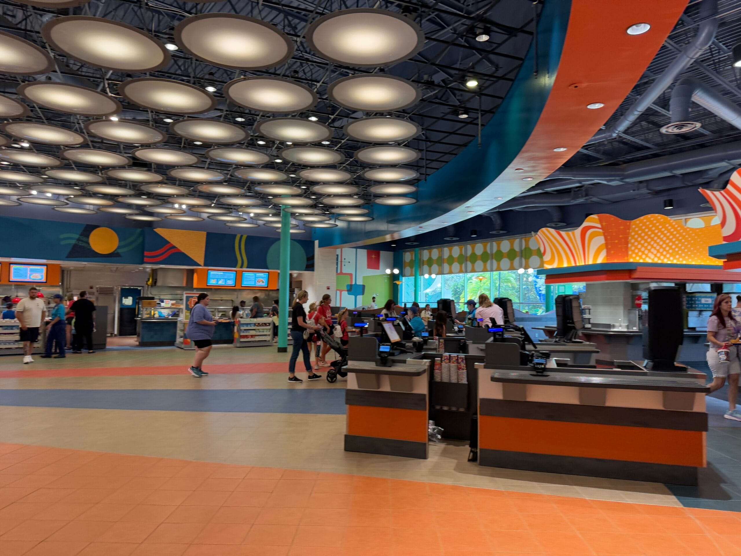 Spacious indoor food court with colorful decor, digital menu boards, and several counters. People stand in line, and circular lights hang from the ceiling.