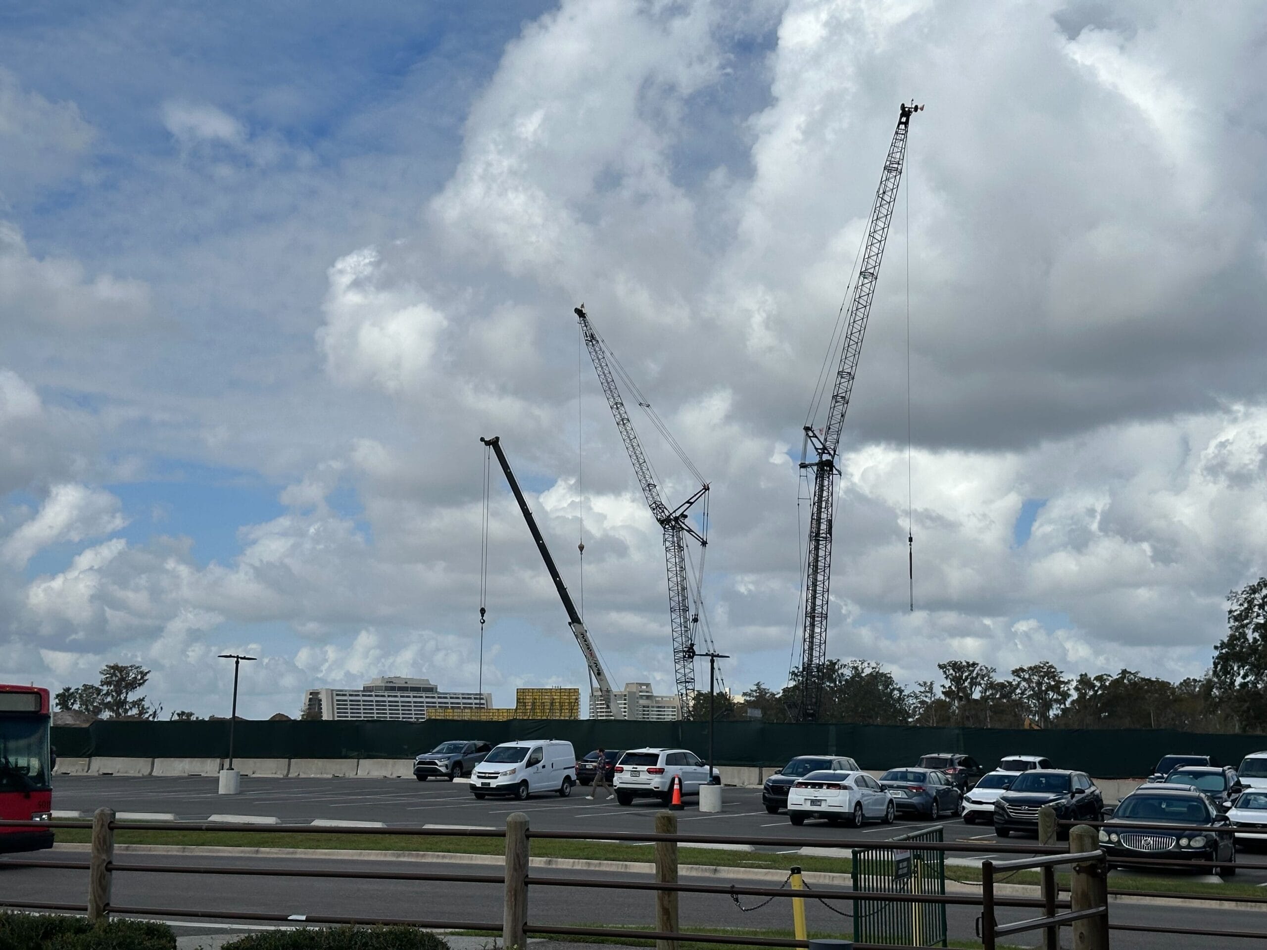 Construction site with three large cranes against a cloudy sky, cars parked in the foreground, and buildings in the background.