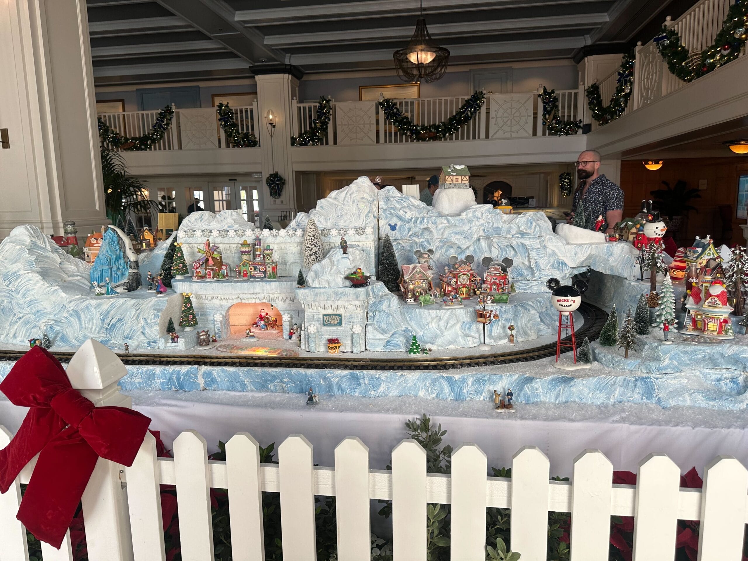 A festive Christmas village display with snowy mountains, decorative houses, and a miniature train set, surrounded by white picket fencing indoors.