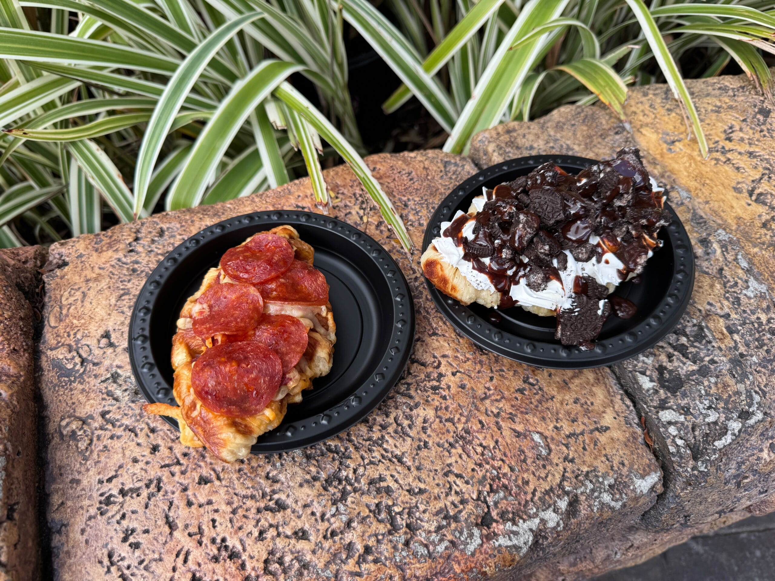 Two black plates on a stone surface: one with a croissant topped with pepperoni, the other with a dessert croissant topped with whipped cream, chocolate sauce, and crumbled cookies.